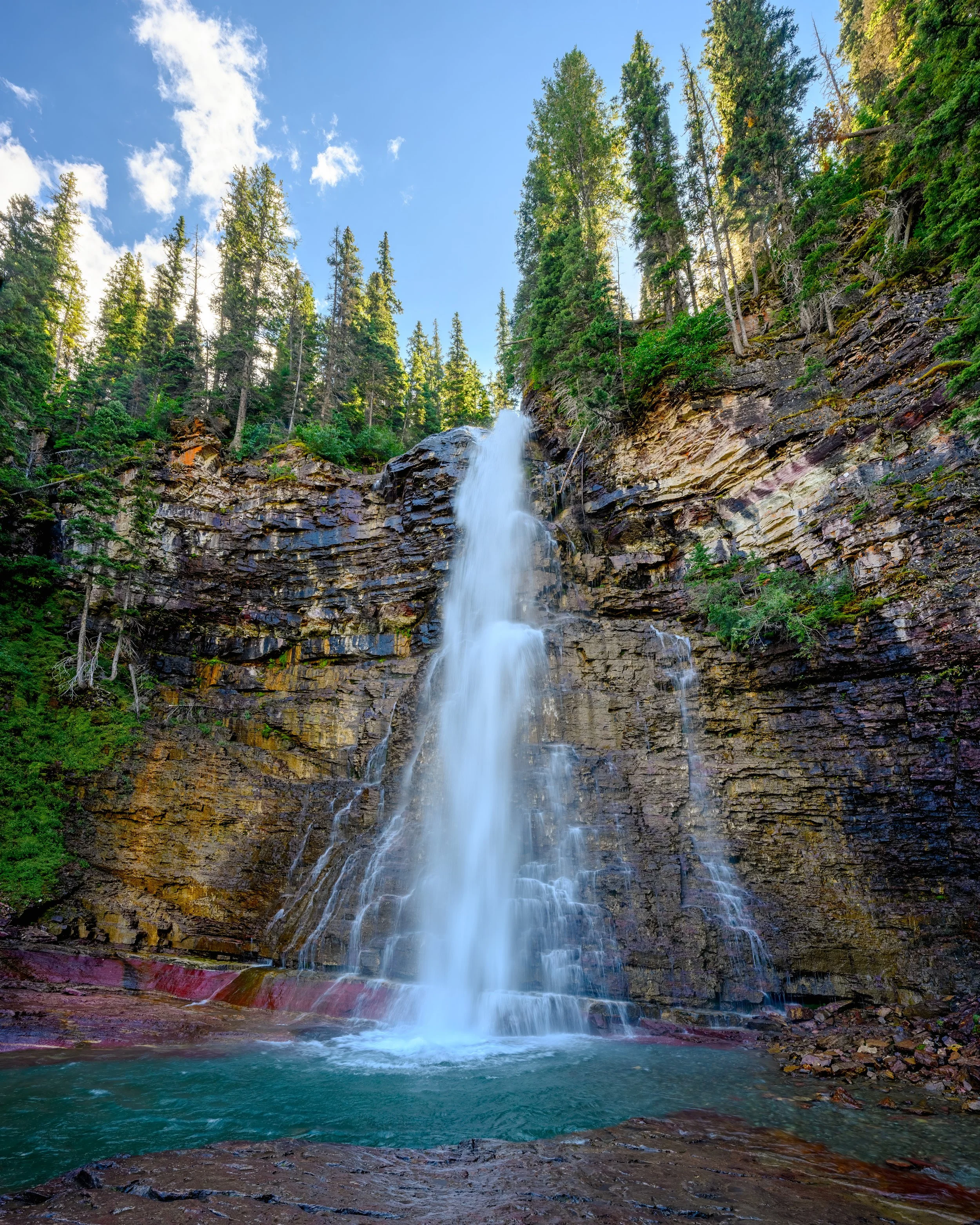 Virginia Falls, Glacier National Park. A waterfall cascading down a rocky cliff into a pool of water, surrounded by tall green pine trees under a blue sky with white clouds.