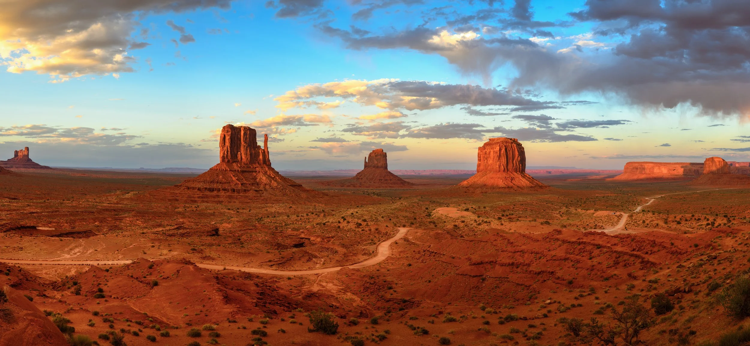 A panoramic view of Monument Valley at sunset with four large sandstone buttes rising from the desert landscape, under a sky with scattered clouds and warm sunlight.