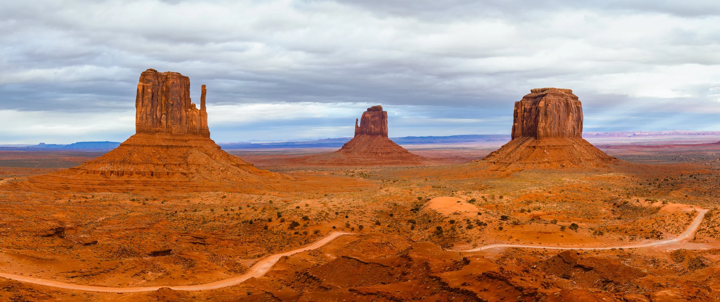 Three large, red sandstone buttes in Monument Valley desert under cloudy sky, with dirt roads winding through arid landscape.