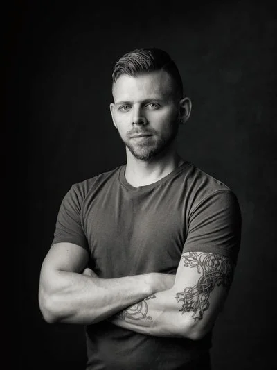 Black and white portrait of a young man with short hair and a beard, wearing a t-shirt, standing with crossed arms against a dark background.