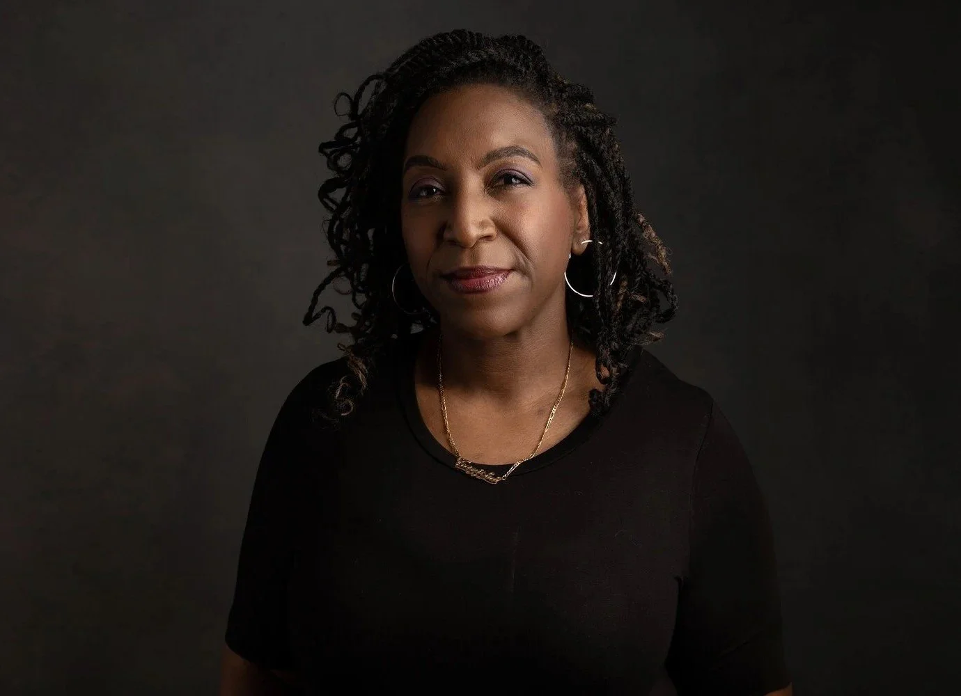 A portrait of a woman with dark curly hair wearing hoop earrings, a necklace, and a black top against a dark background.
