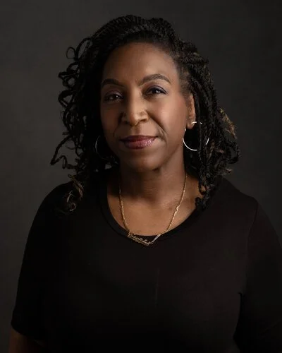 A woman with curly hair wearing hoop earrings and a black top, posing against a dark background.