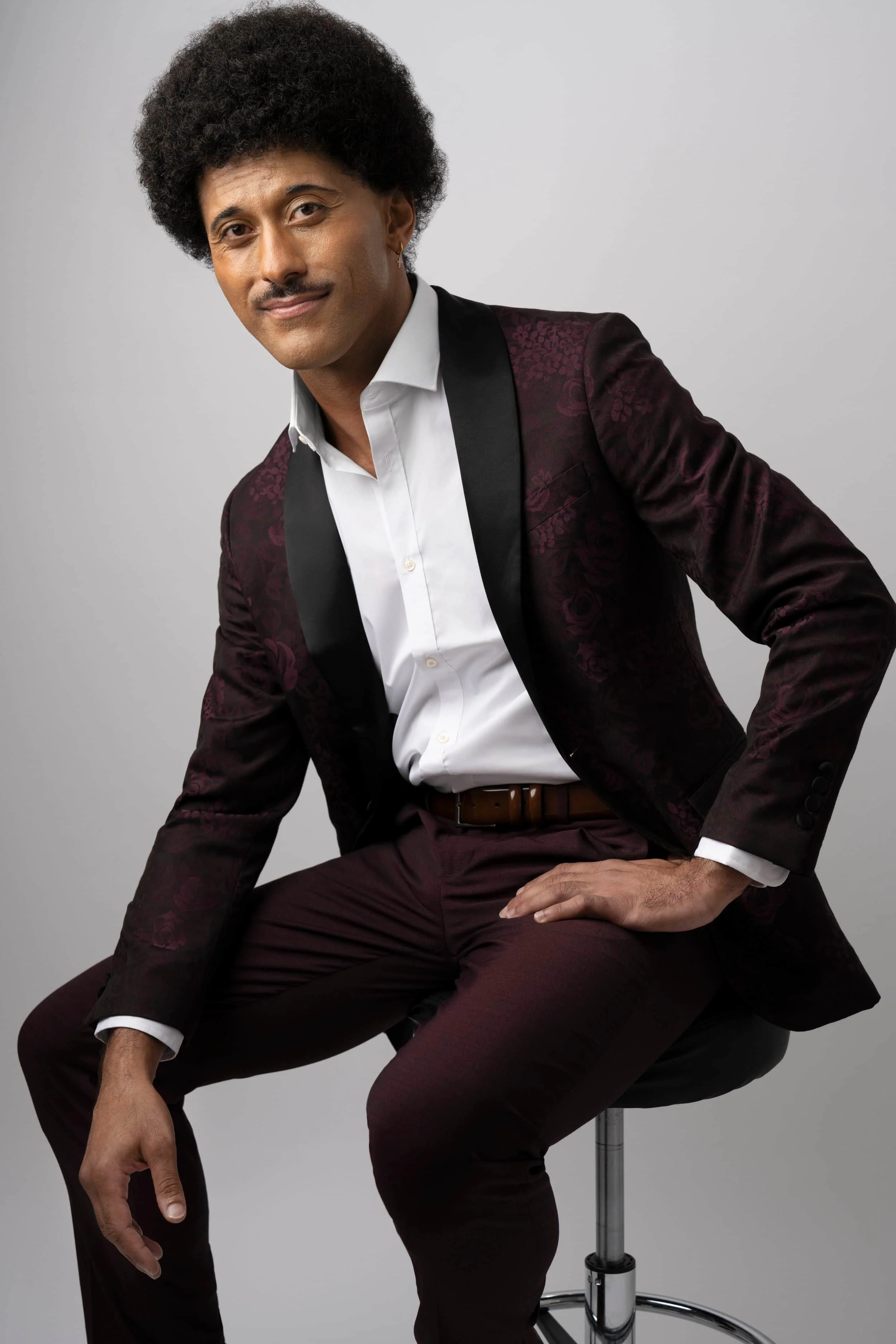 A man with an afro hairstyle, wearing a white dress shirt, a dark red patterned blazer, and matching pants, sitting on a stool with one hand on his knee against a plain gray background.