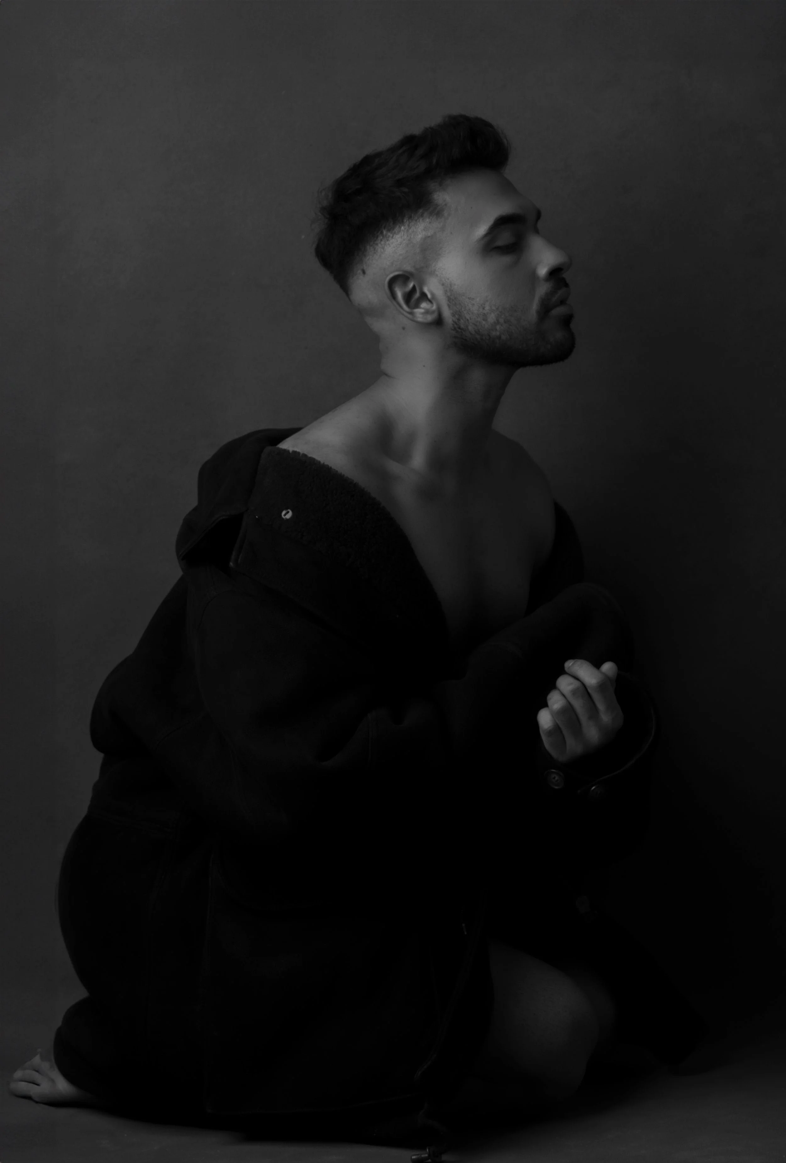 A black and white photo of a shirtless man sitting on the floor, wearing a dark jacket draped over his shoulders, with his eyes closed and head tilted slightly upward.