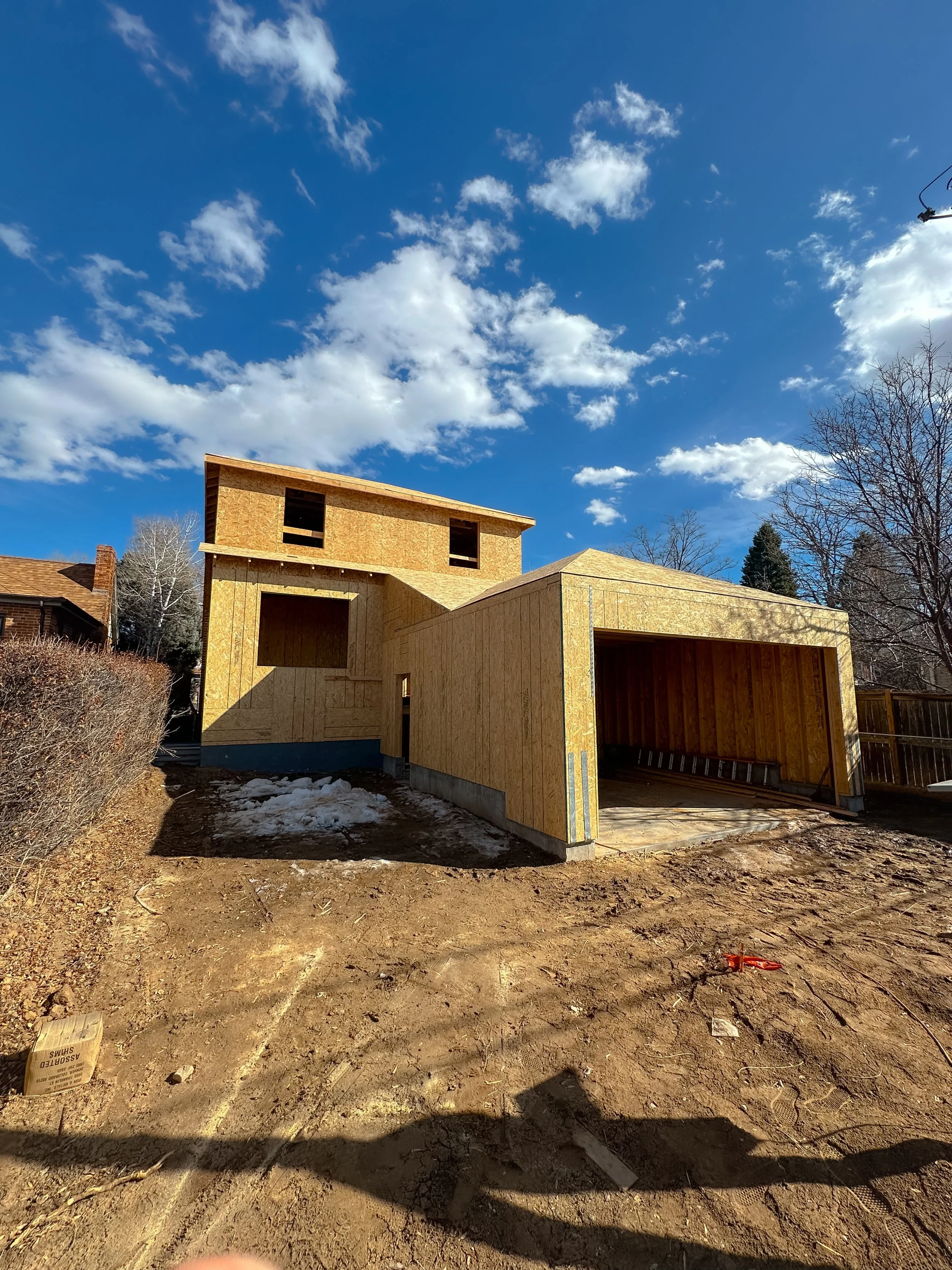 A house under construction with wooden framing, a garage, and clear blue sky with scattered clouds.