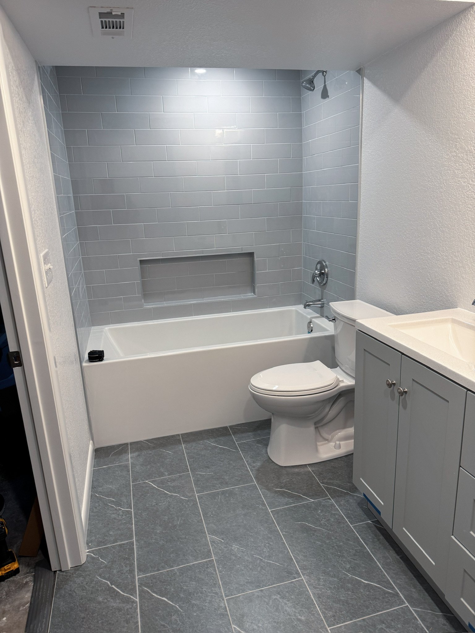 Bathroom with gray tile walls and floor, a bathtub, a toilet, gray cabinetry with a sink, and a built-in shower niche