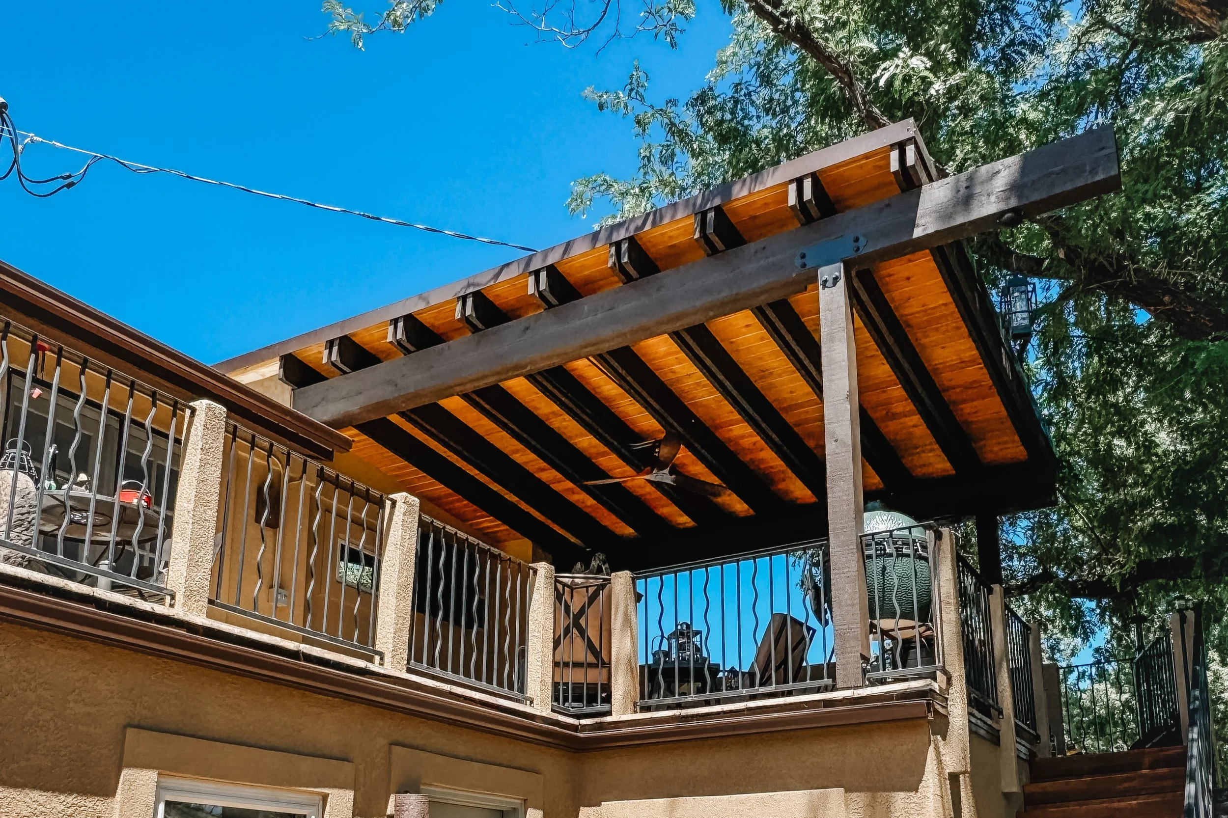View of an outdoor balcony or patio with a pergola or roof structure made of wood and metal, decorated with string lights, and surrounded by a metal railing. The sky in the background is bright blue with some trees visible.