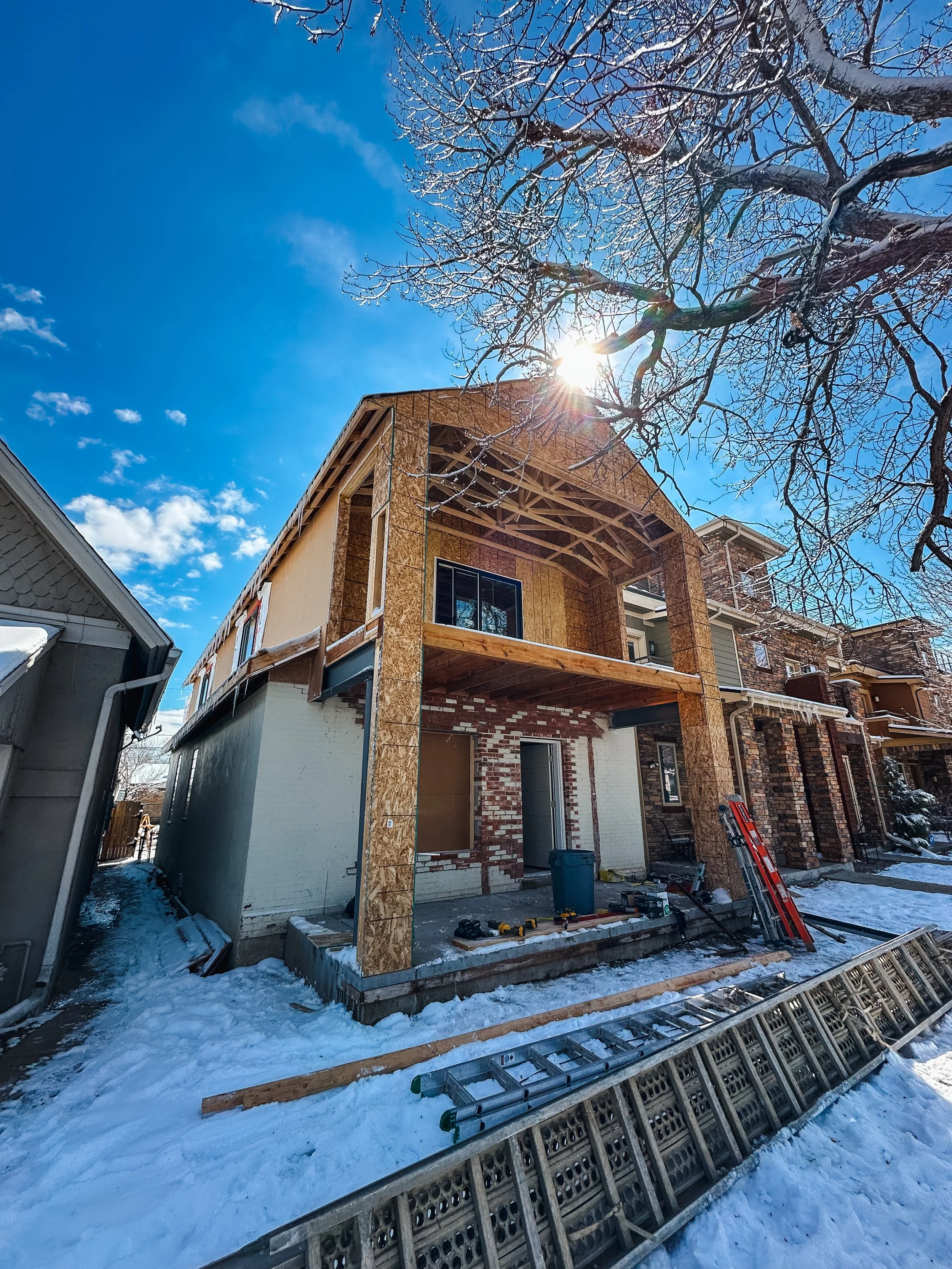 A house under construction with exposed wooden framing and partial brick wall, surrounded by snow in a residential neighborhood