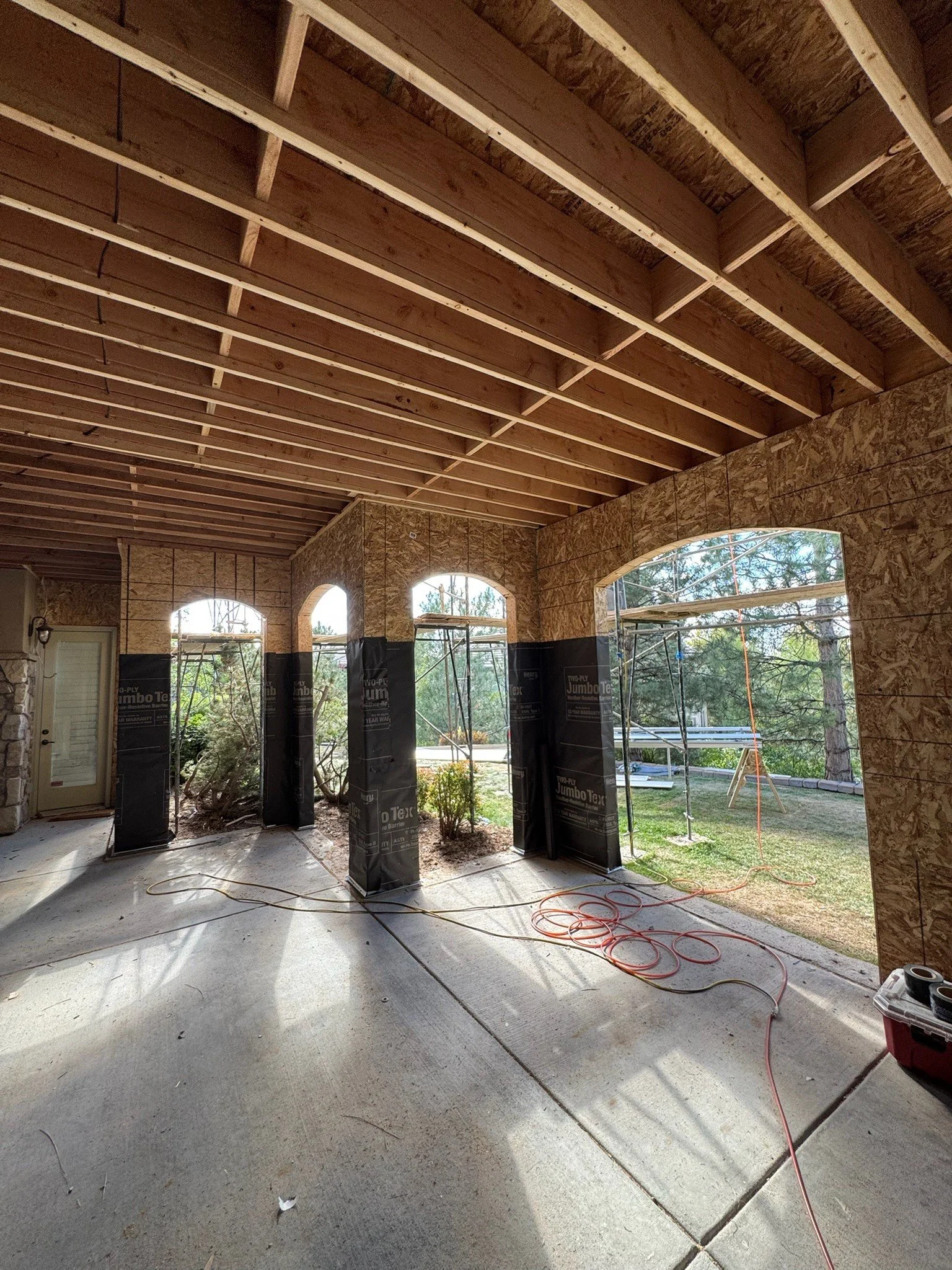 Interior of a house under construction with unfinished walls and ceiling, showing wooden framing and large arched windows, with construction materials and tools on the floor.