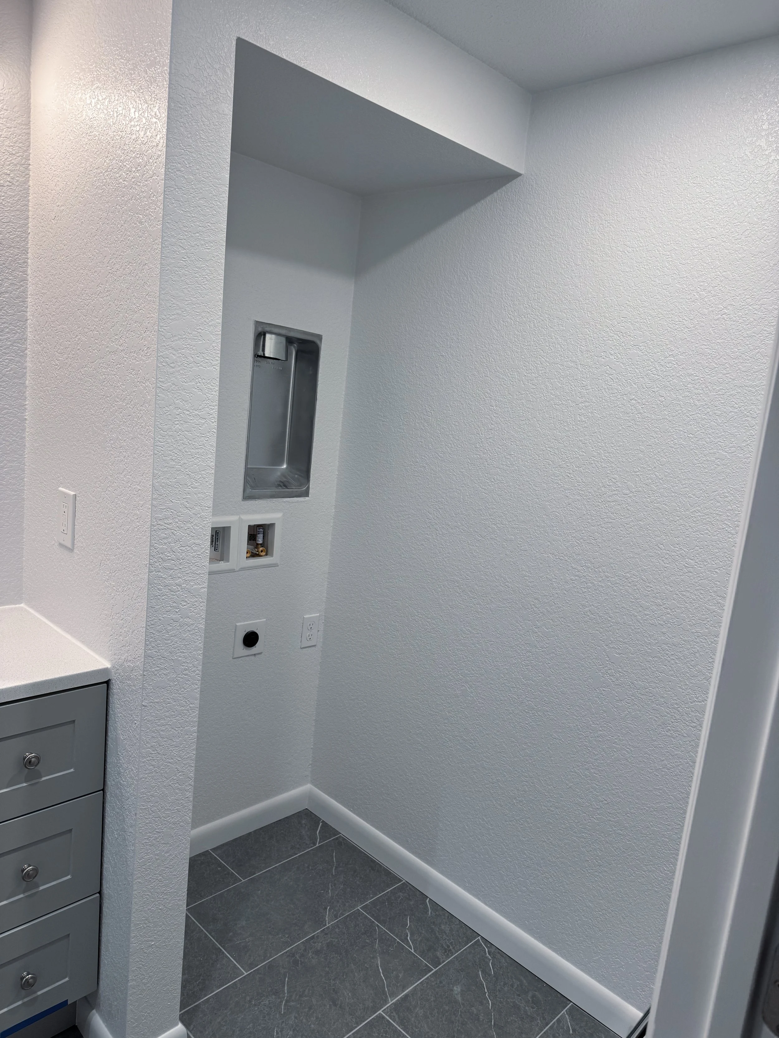 Empty laundry room with textured white walls, black tiled floor, and a built-in soap dispenser. There are electrical outlets and a small gray cabinet.