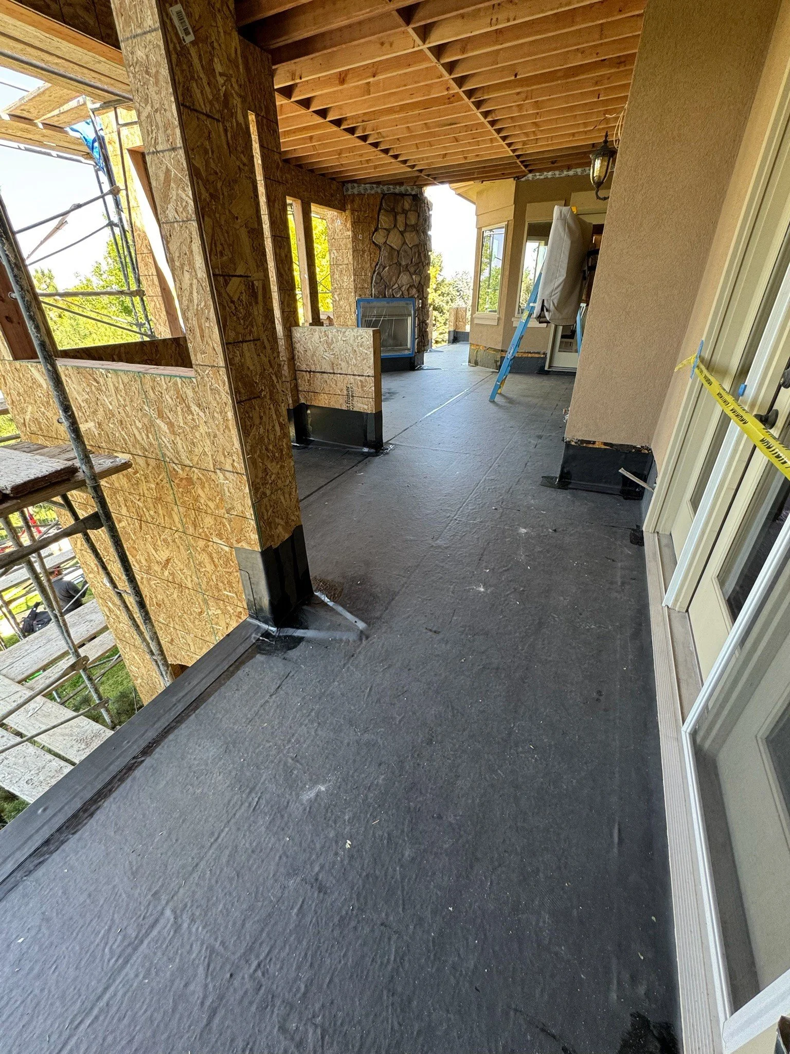 Construction site on a balcony with exposed wooden ceiling and partially built wall, a ladder, and a clear view of tree and sky outside.