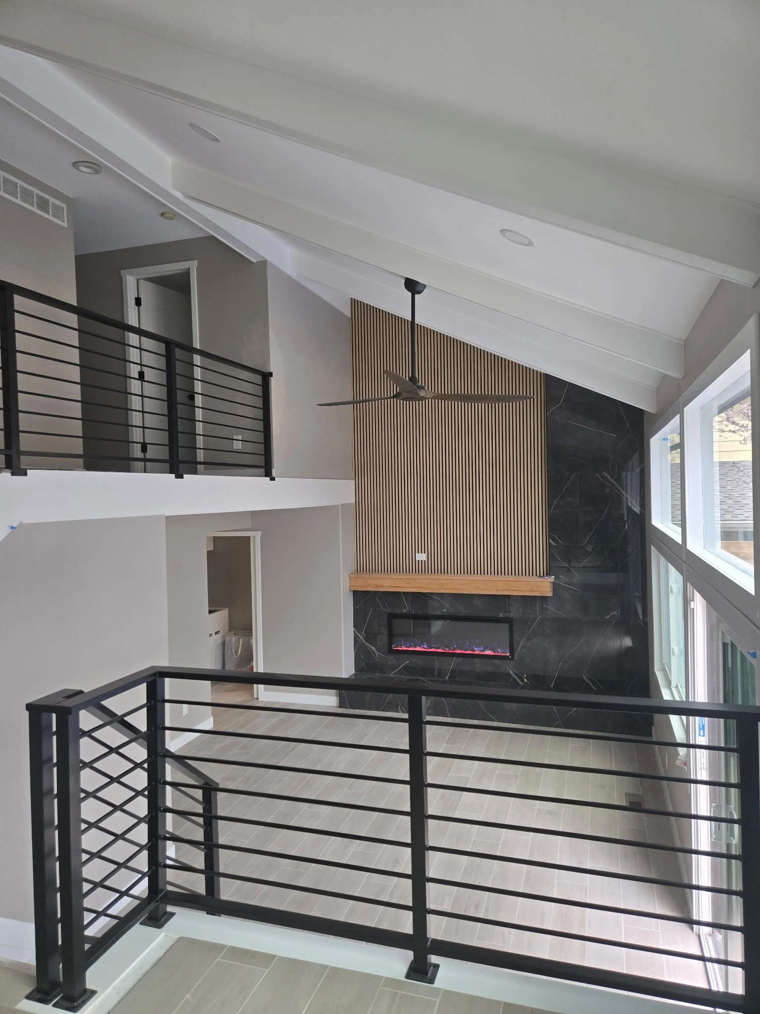 Interior view of a modern home with an open second floor, black metal stair railing, wooden accent wall, fireplace, large windows, and a ceiling fan.