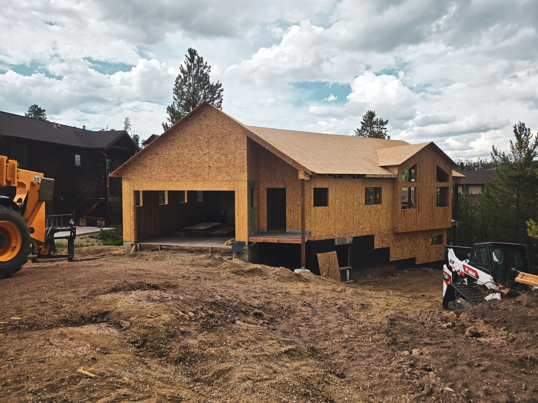 Construction site with a two-story house under construction, made of wood panels with an open garage and multiple windows, surrounded by construction equipment and dirt ground, in a neighborhood with trees and other houses nearby.