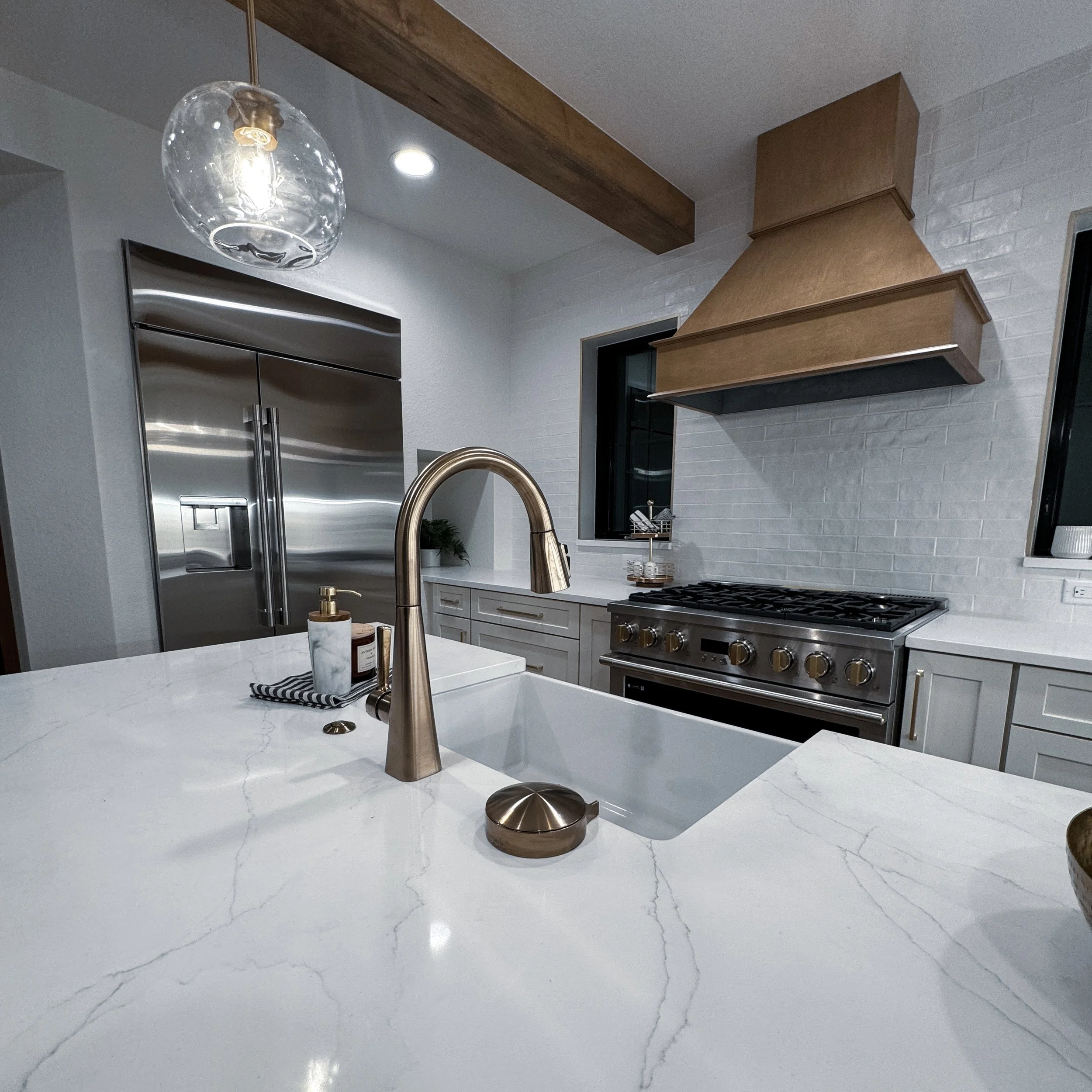 Modern kitchen with marble countertop, gold faucet, stainless steel refrigerator, beige range hood, and white brick backsplash.