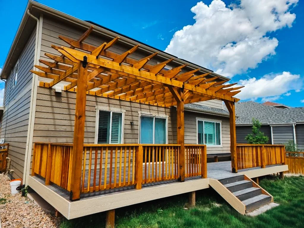 Wooden deck with a pergola attached to a beige house with white trim, against a blue sky with white clouds.
