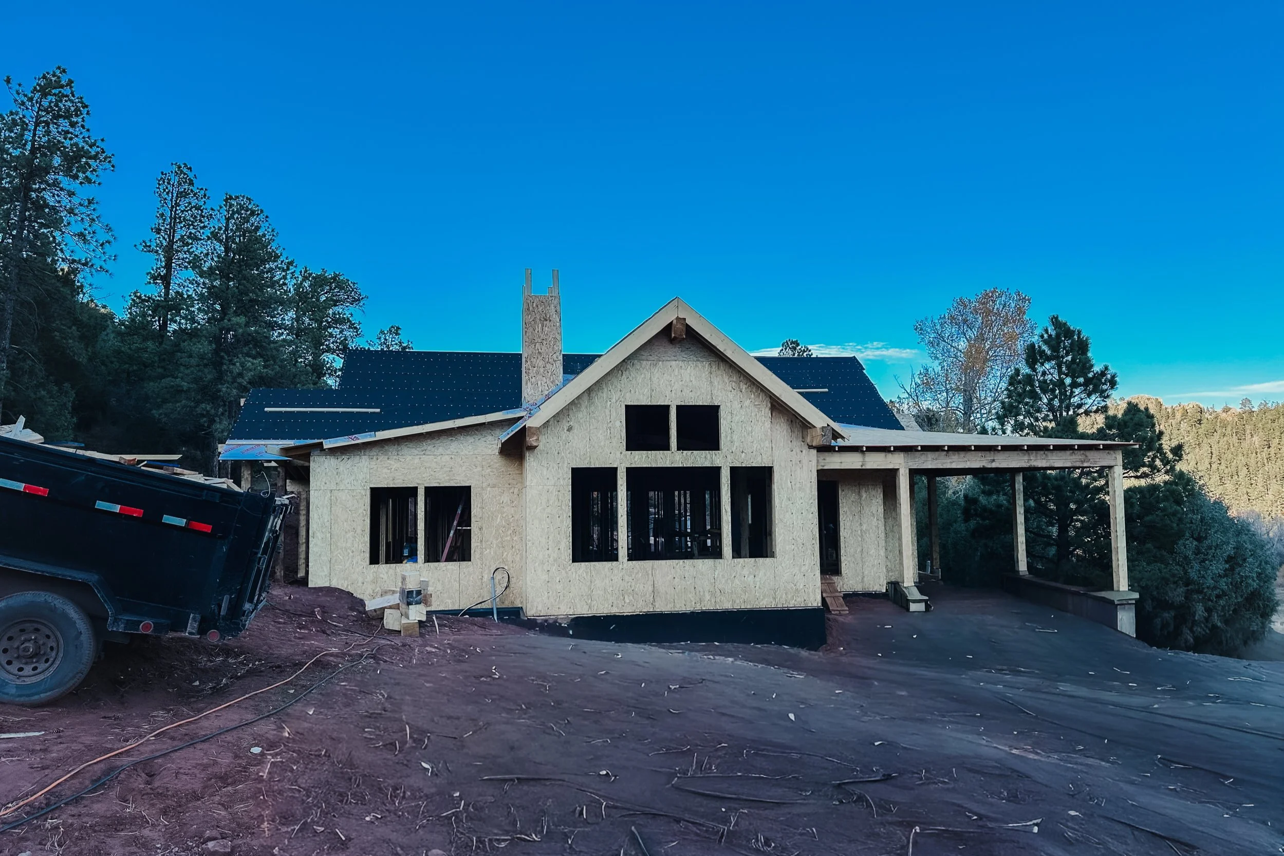 A house under construction with unfinished exterior walls, black window frames, and a black roof, surrounded by trees and dirt ground, under a clear blue sky.