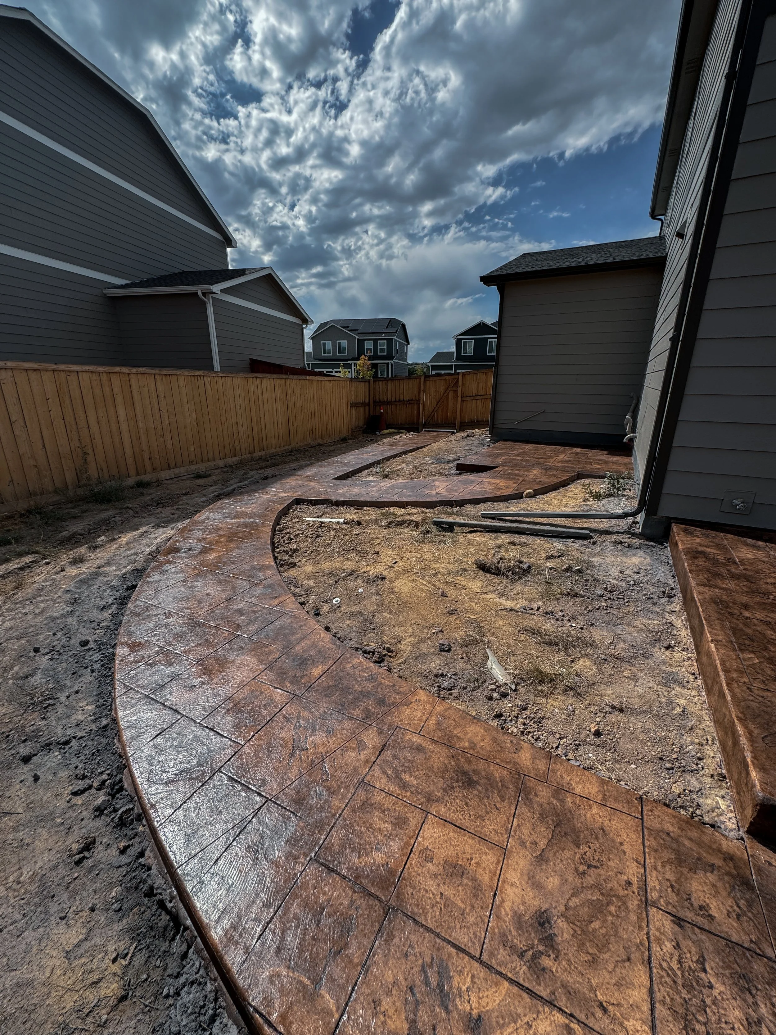 Backyard with newly installed curved brick pathway, surrounded by dirt and construction materials, with a wooden fence and neighboring houses under cloudy sky.