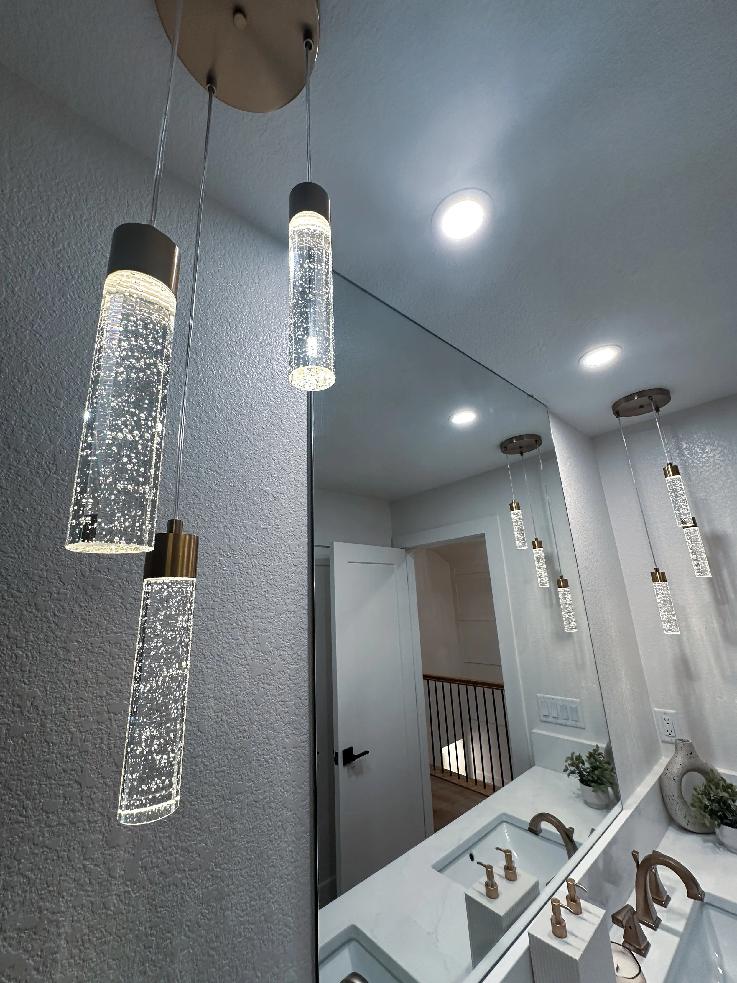 Bathroom mirror with multiple pendant lights hanging from ceiling and a white countertop with two sinks, fixtures, and decorative plants.