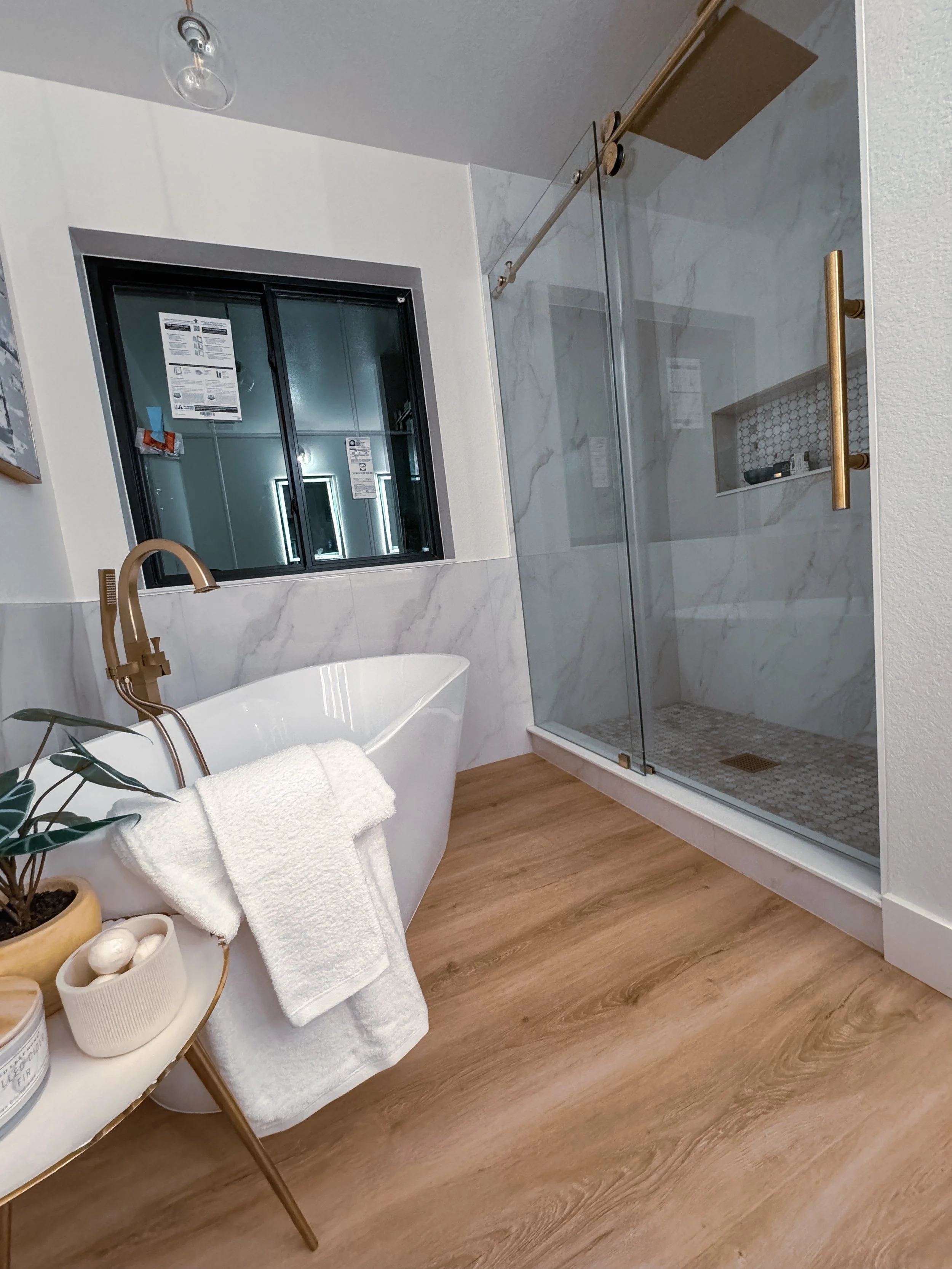 Modern bathroom with a freestanding bathtub, a potted plant, a draped white towel, a glass shower with gold fixtures, a window, and wood flooring.