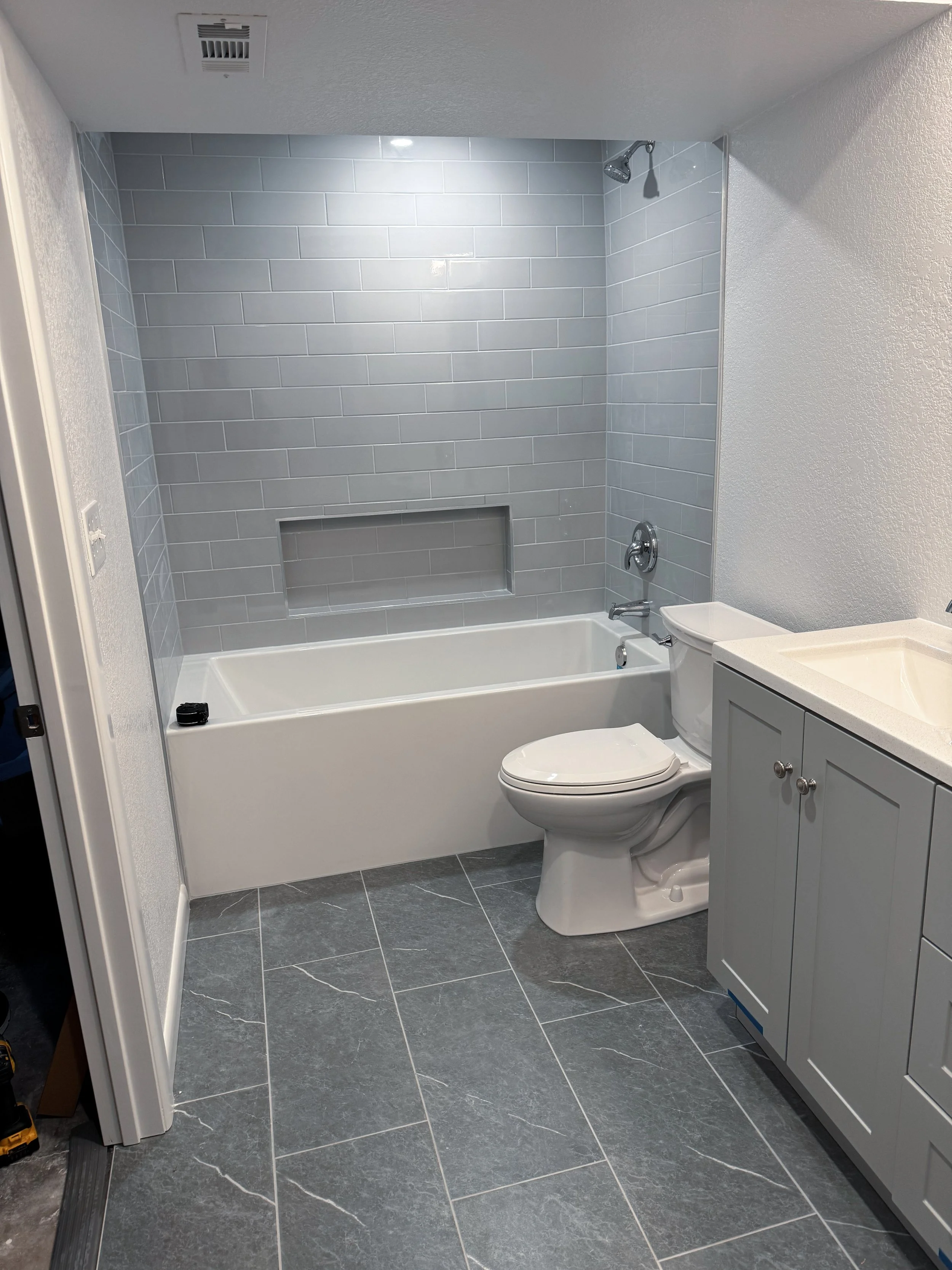 A modern bathroom with a bathtub, a toilet, gray tile walls, and a white vanity with a sink.