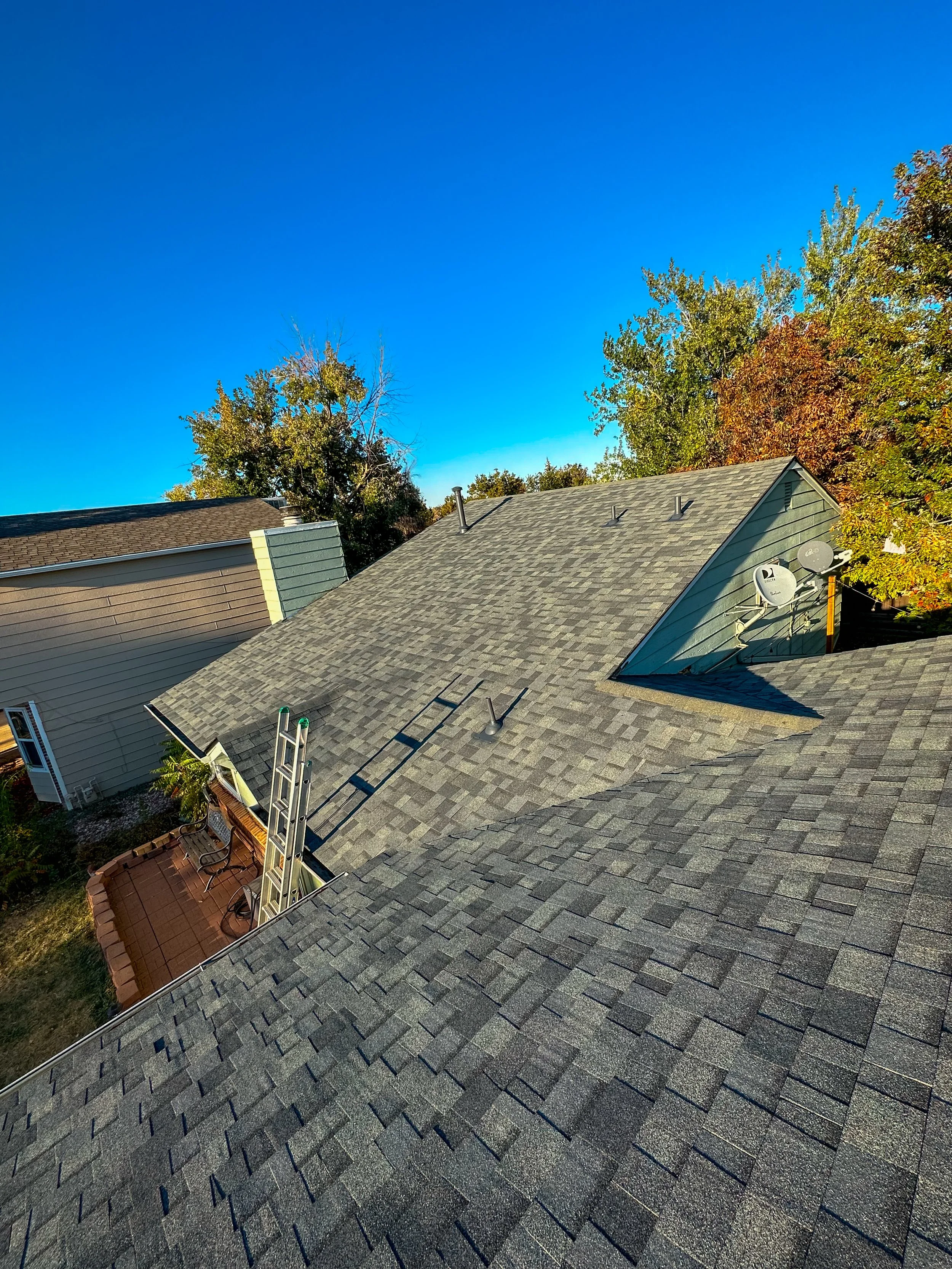 Aerial view of a house roof with gray shingles, satellite dishes, and a ladder leaning against the roof, with trees and clear blue sky in the background.