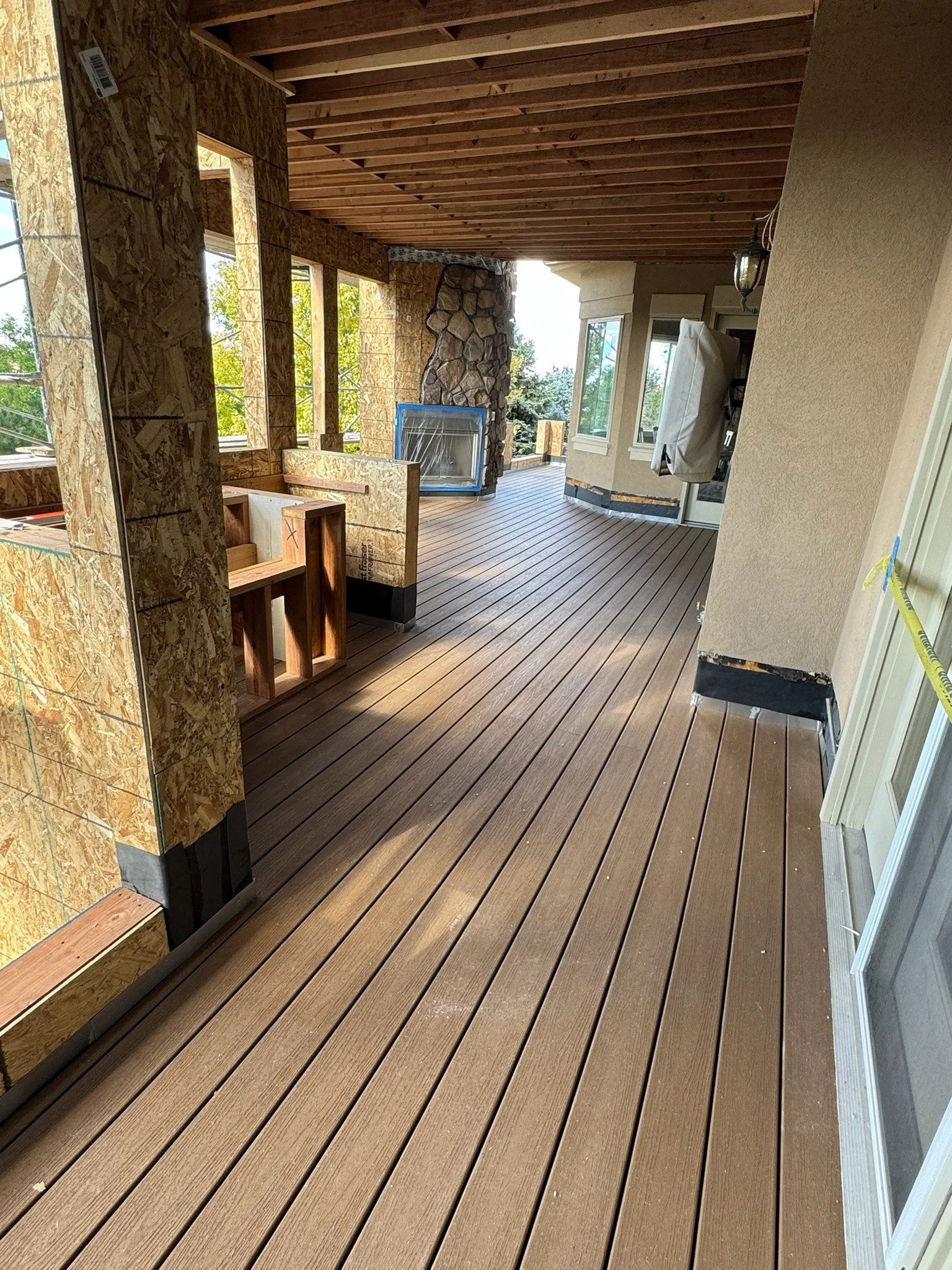 A house porch under construction with new wooden flooring and walls made of oriented strand board (OSB); some windows are installed, and other building materials are present.