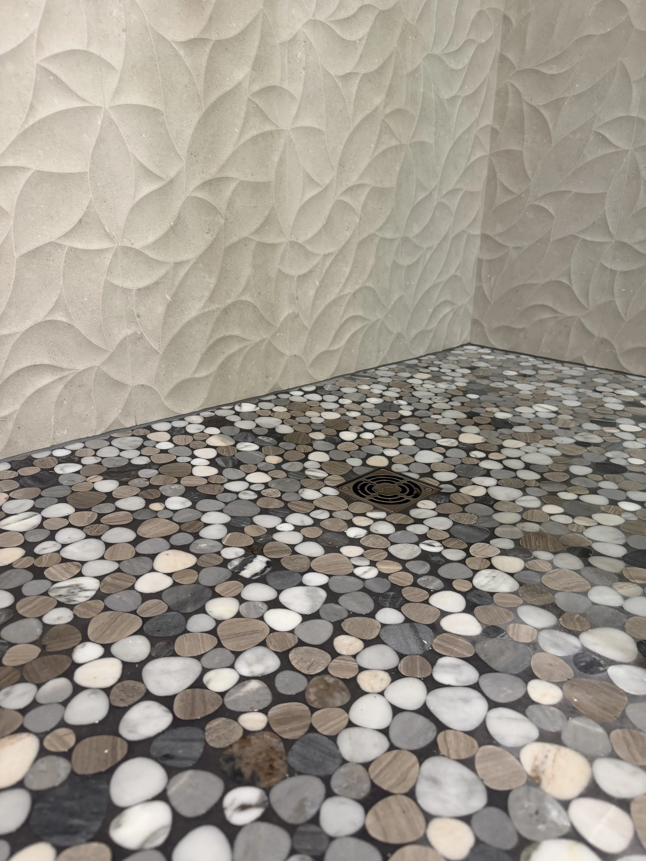 Close-up of a textured beige wall with three-dimensional leaf-shaped patterns and a floor made of small, rounded multicolored stones surrounding a metal drain.