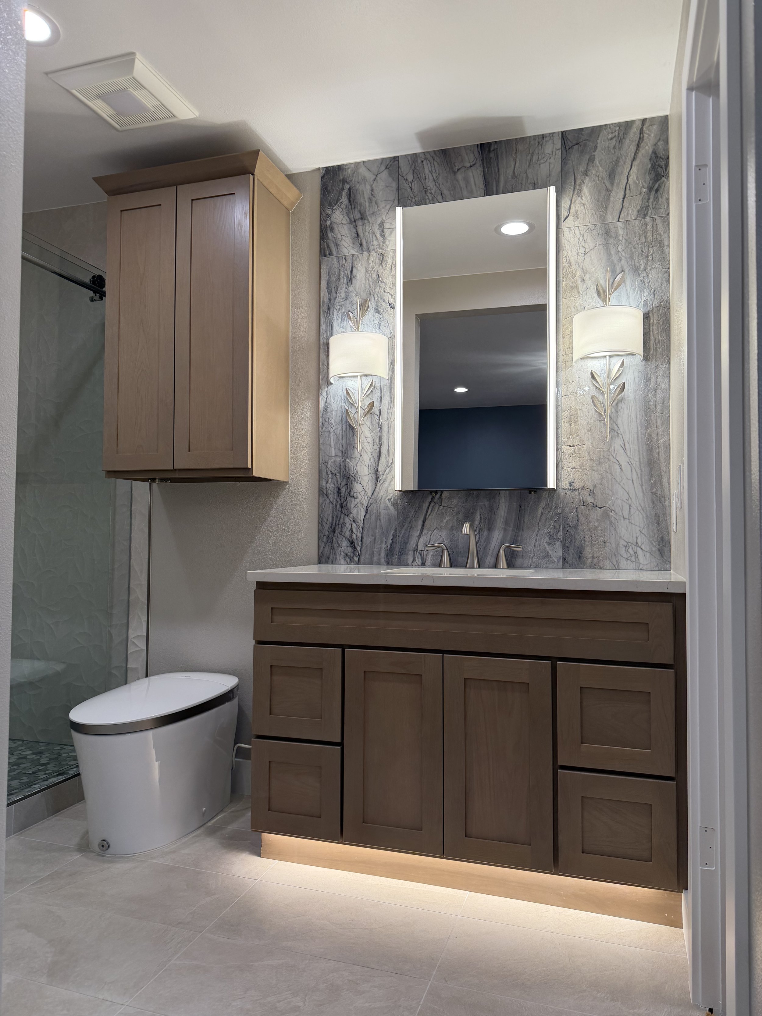 Bathroom with wooden vanity, marble backsplash, mirror, wall sconces, and modern toilet.