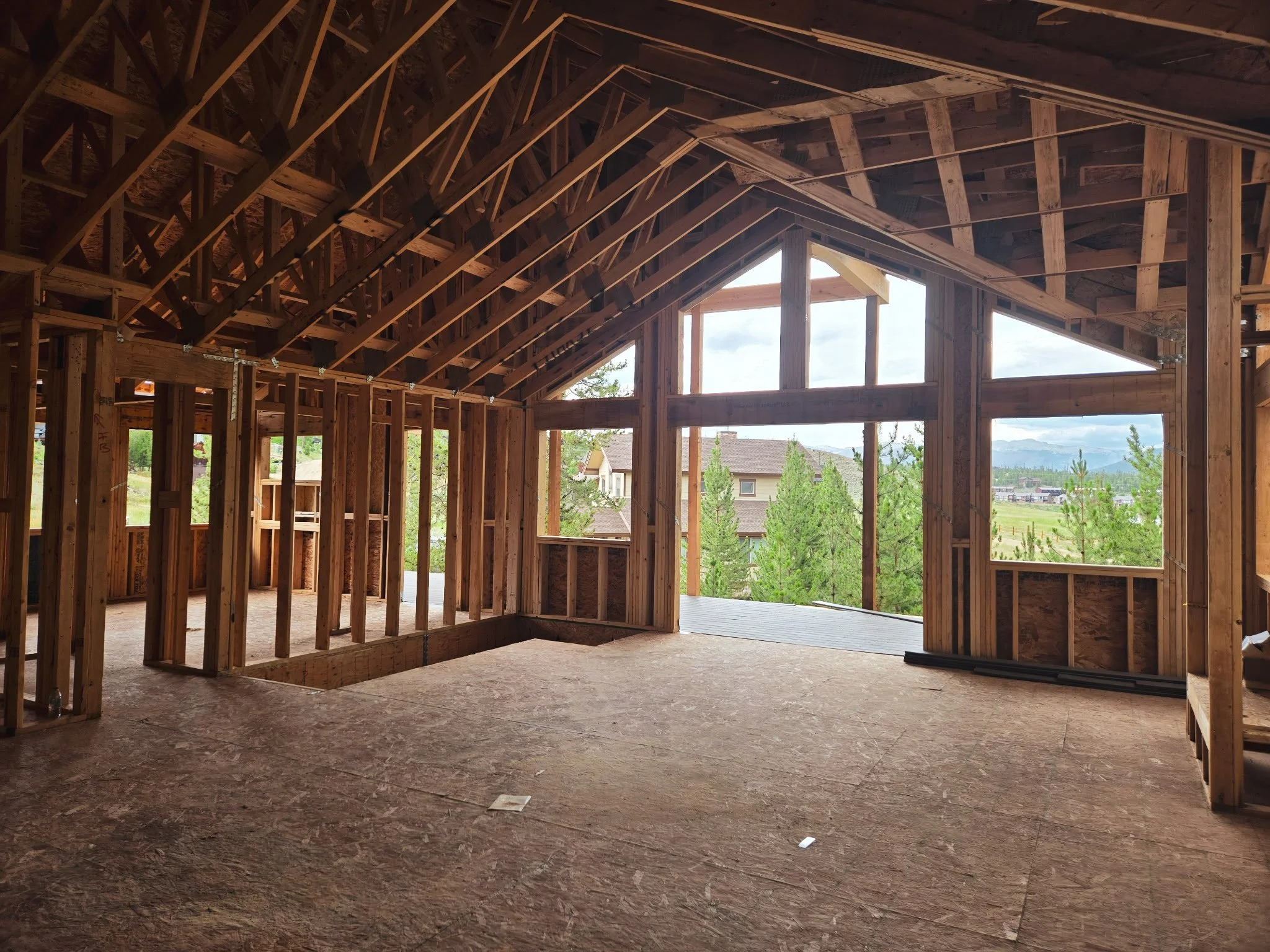 Interior view of a house under construction with exposed wooden framing, open windows, and a view of trees and mountains outside.