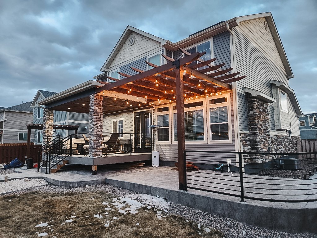 Backyard of a modern two-story house with a covered deck, string lights, patio furniture, and a newly landscaped yard.