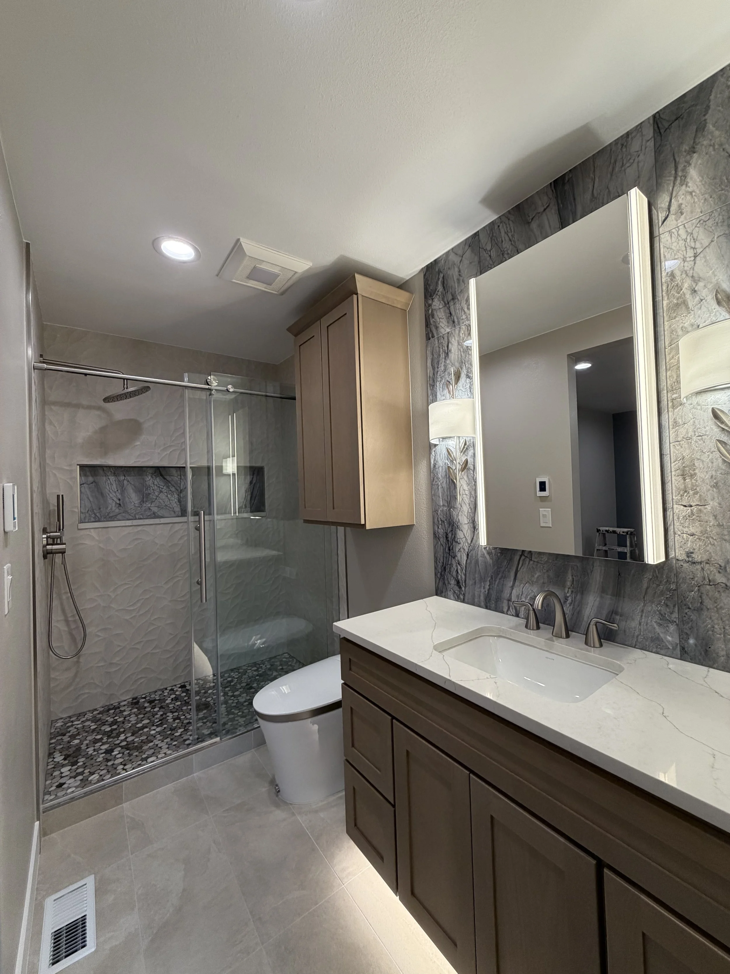 Bathroom with a vanity mirror, marble wall, wooden cabinets, marble countertop, sink, faucet, walk-in shower with glass door, rain shower head, white toilet, beige floor tiles, and a ceiling vent.