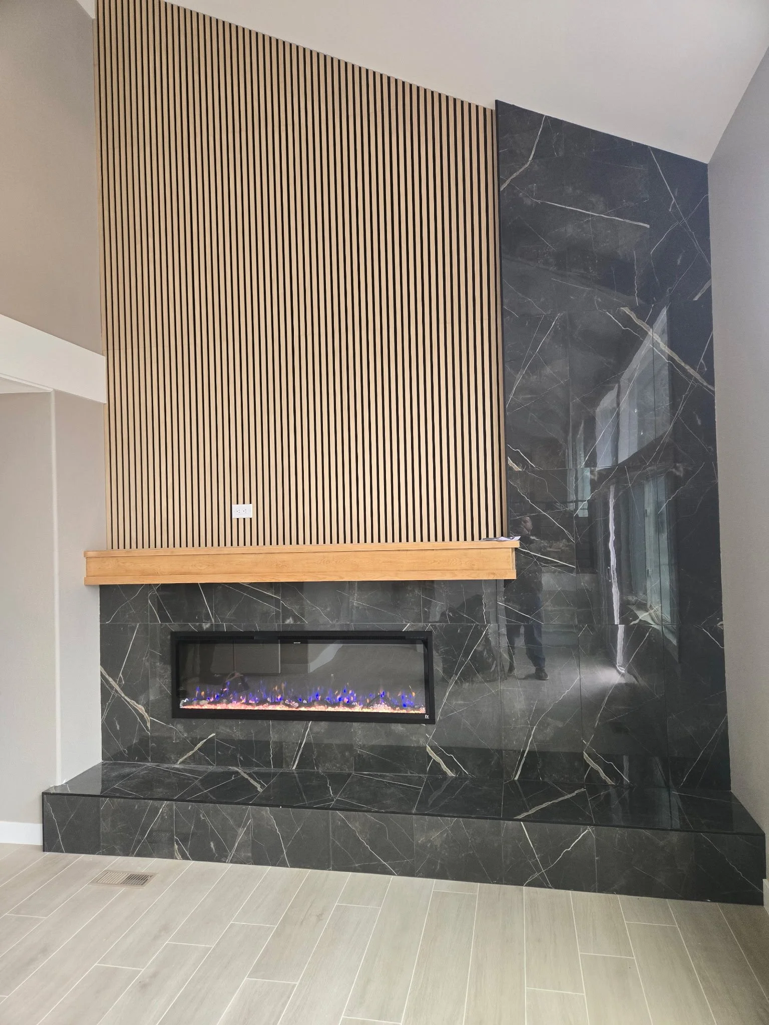 Modern living room corner with black marble wall, electric fireplace, wooden slat panel, and gray floor tiles.