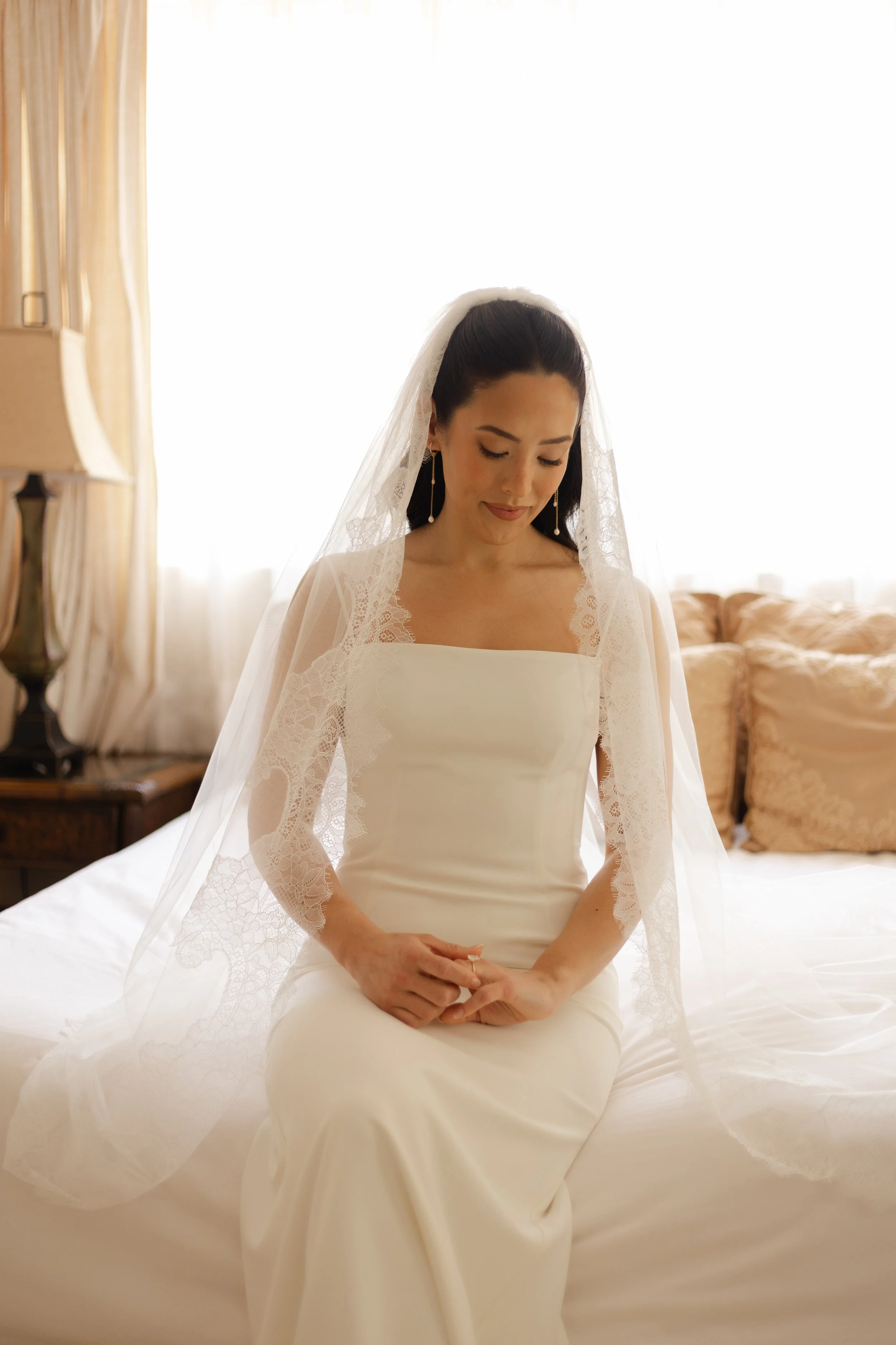 A bride in a white wedding dress with lace sleeves sitting on a bed with a soft expression.