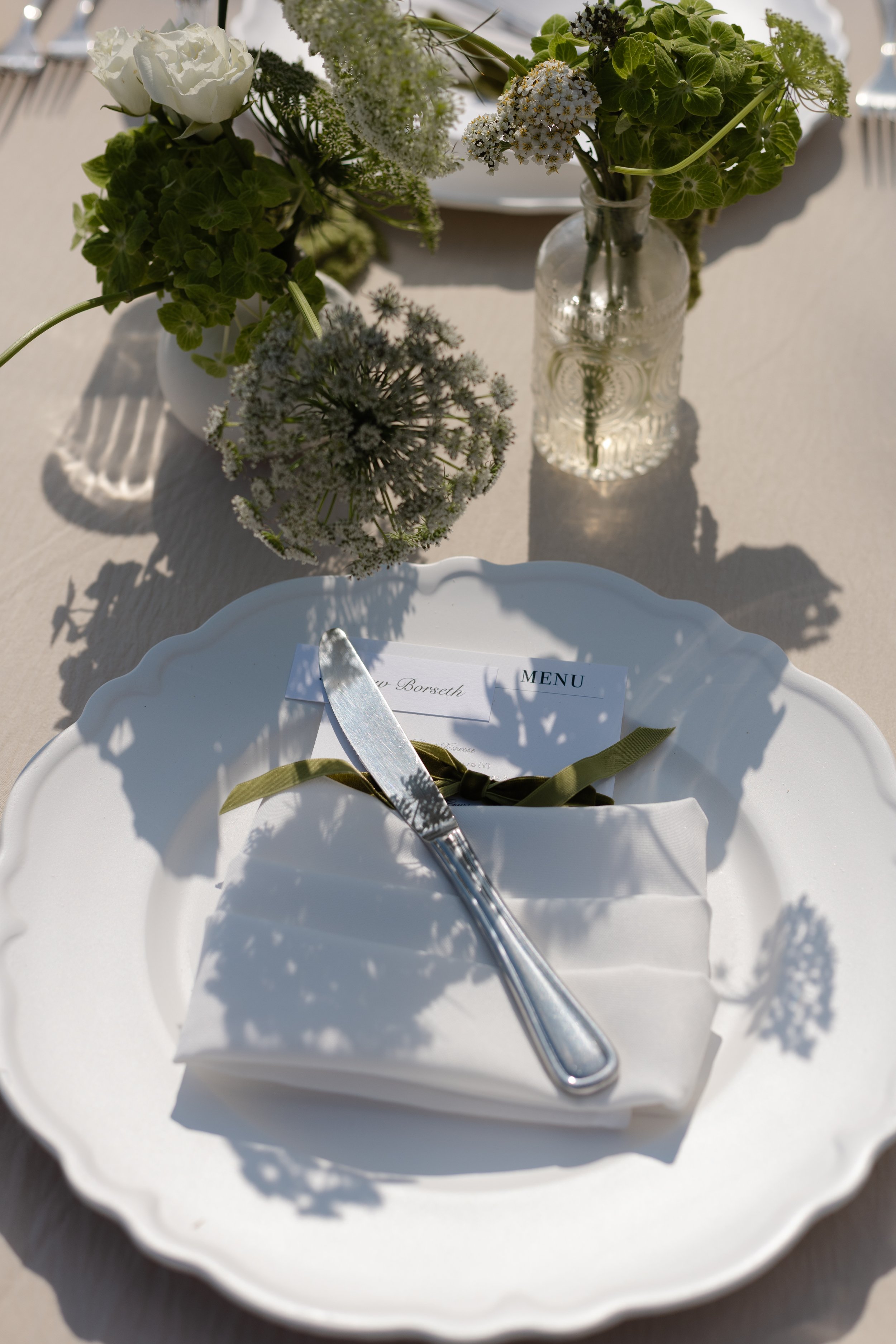 A table setting with a white plate, a folded napkin with a green ribbon, a knife, a menu, and floral centerpieces in vases filled with white and green flowers, all illuminated by sunlight casting shadows.