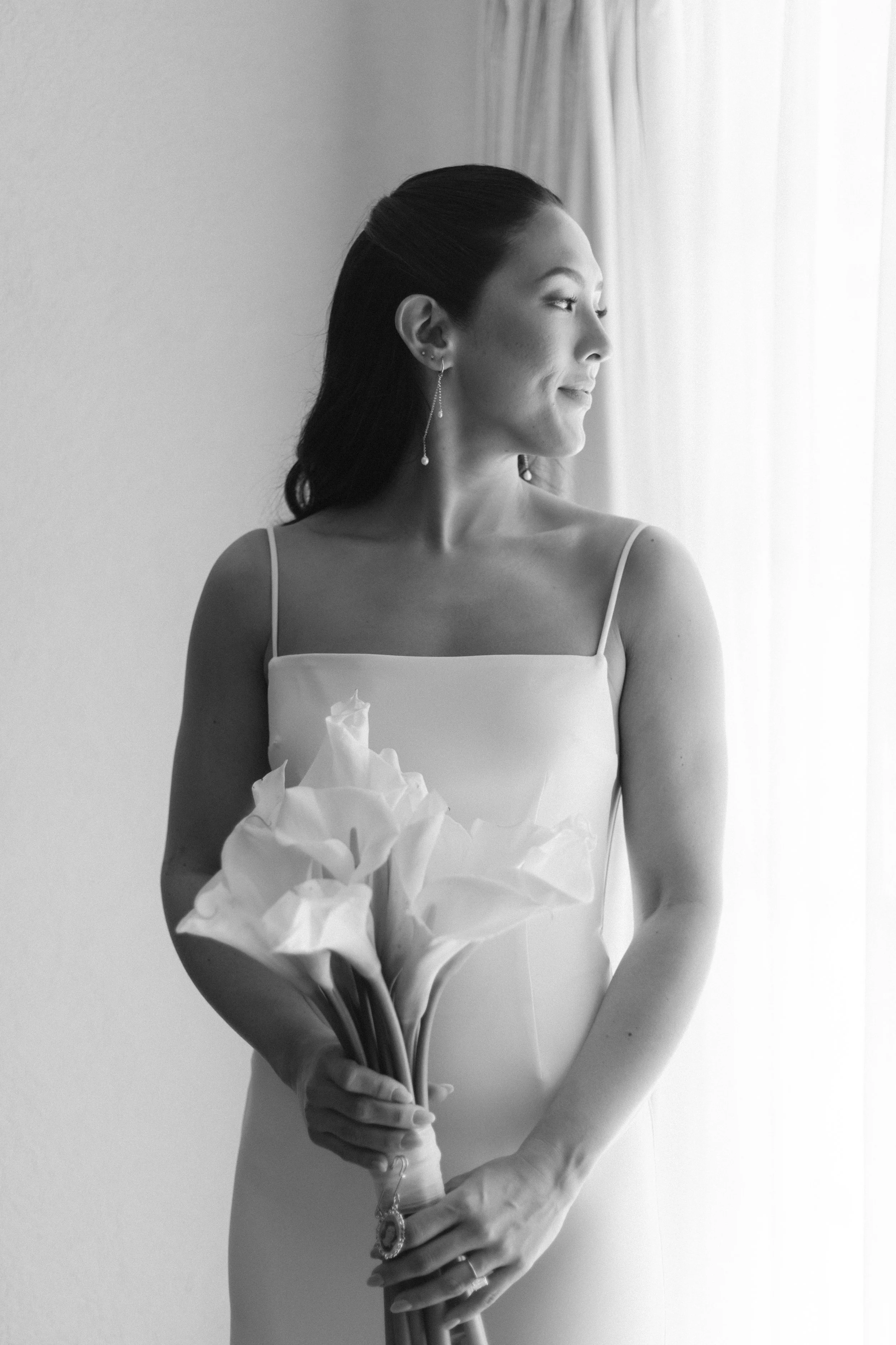 A woman in a wedding dress holding a bouquet of calla lilies, standing near a window with curtains, looking out.
