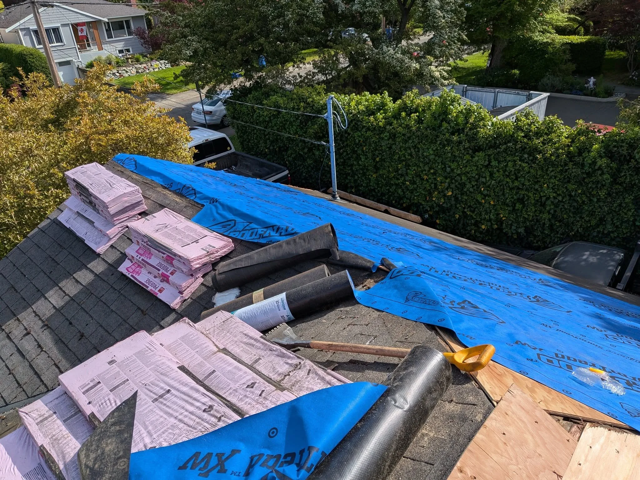 Roof under construction with pink insulation, black roofing underlayment, and blue weather-resistant barrier, with trees, parked cars, and residential buildings in the background.