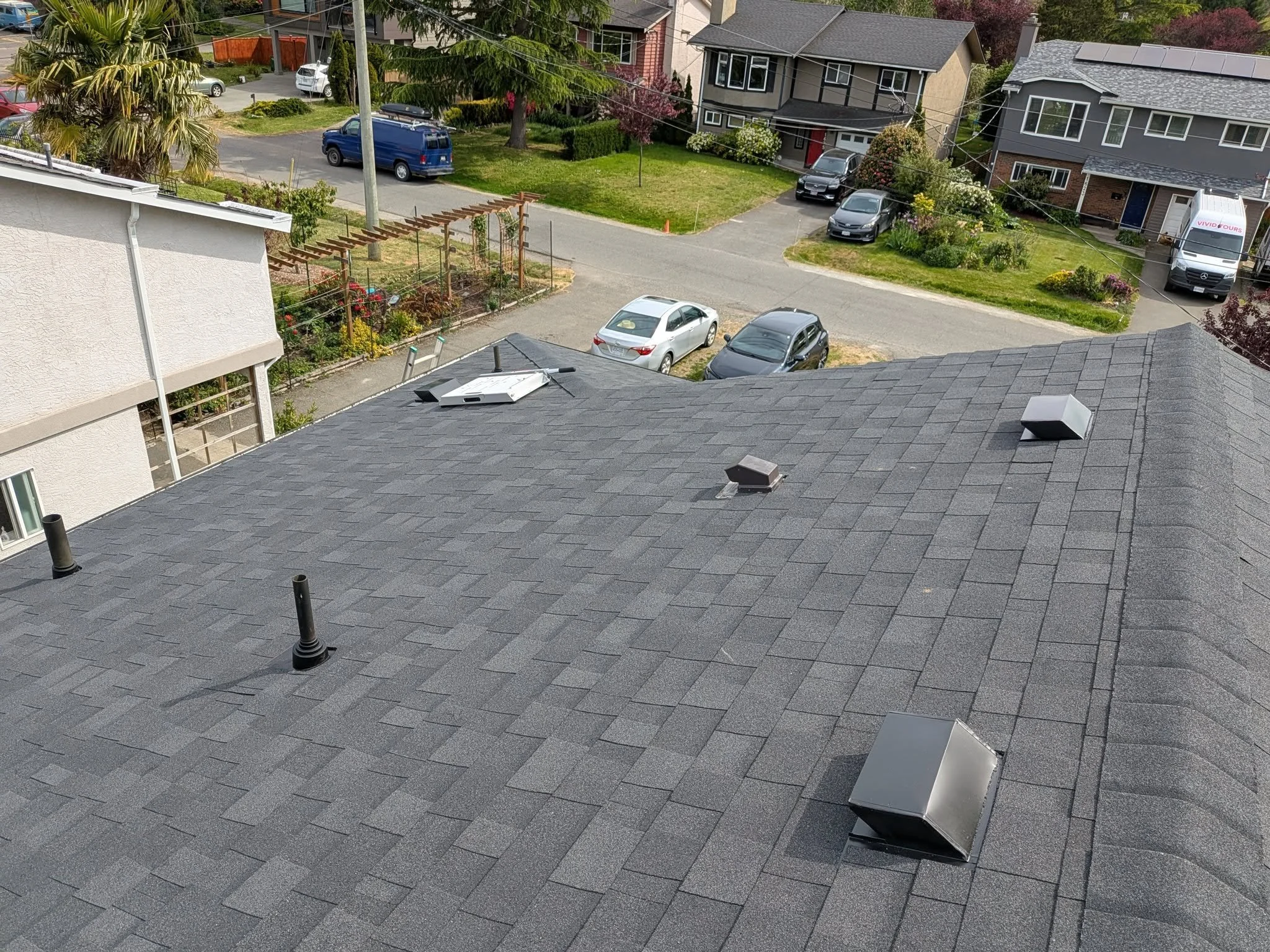 View of a residential rooftop with multiple vents and skylights overlooking a suburban neighborhood with parked cars, houses, and gardens.