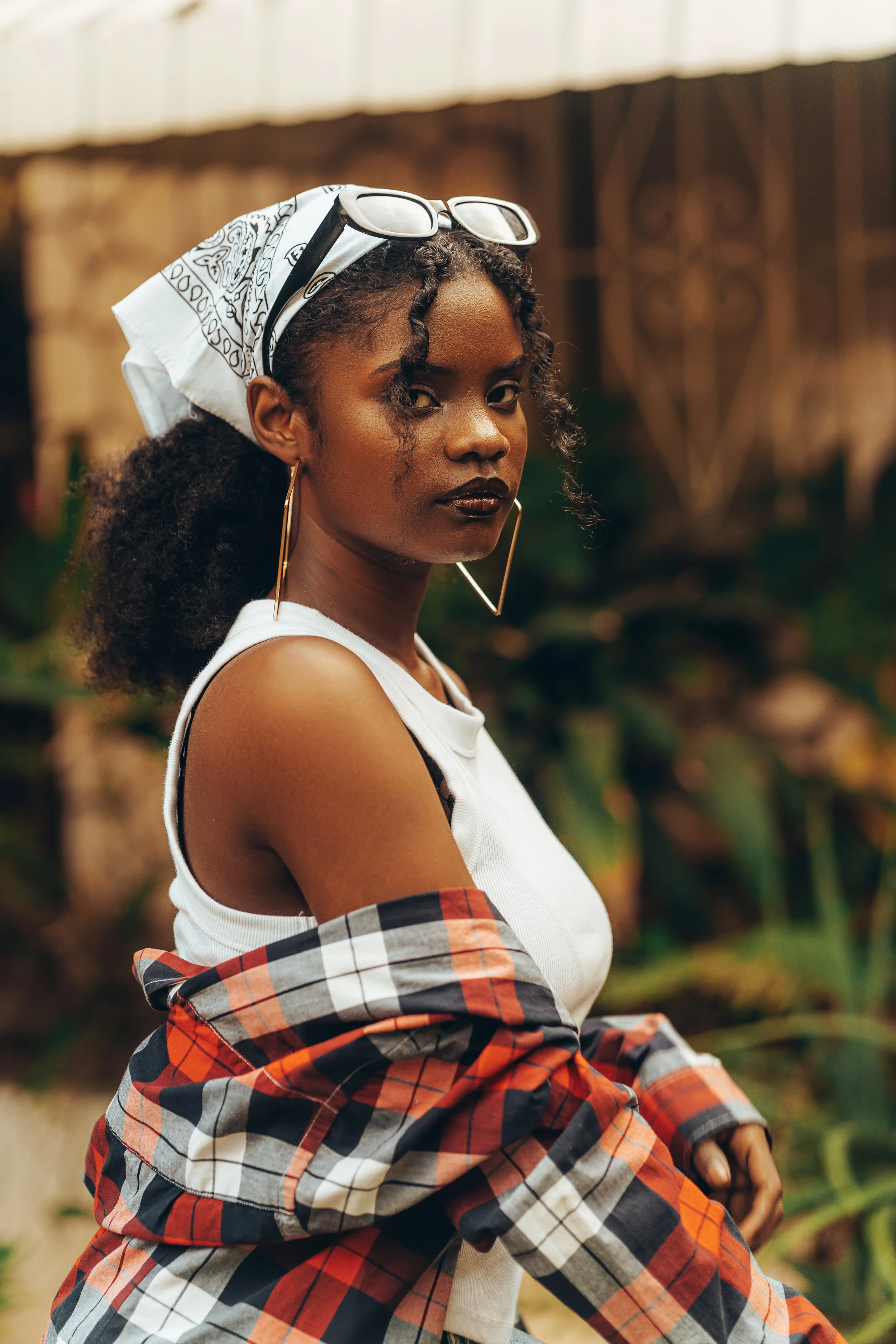 A woman with dark, curly hair, wearing sunglasses on her head, gold hoop earrings, dark lipstick, a white tank top, and a plaid shirt around her waist, sitting outdoors with blurred plants in the background.