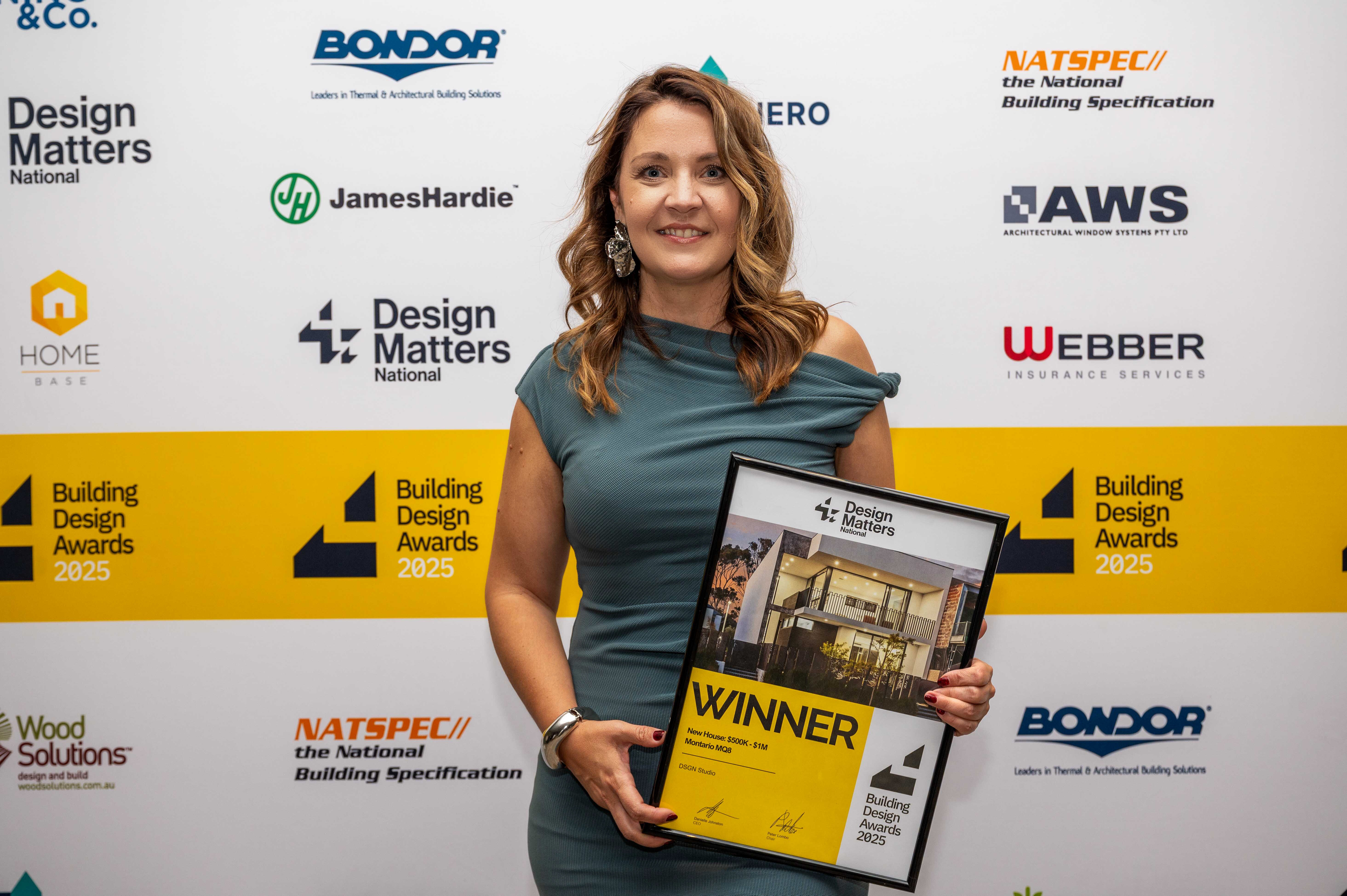 Danica Novakovic, wearing a teal dress is holding a framed award certificate that says 'WINNER' for Design Matters building project award, standing in front of a backdrop with logos and text related to a building design awards event.