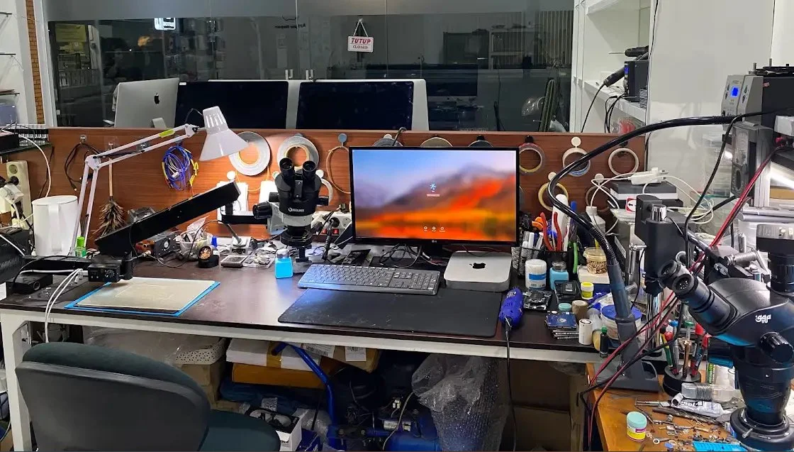 Messy science or electronics workbench with microscopes, computers, tools, and various electronic components and lab equipment.