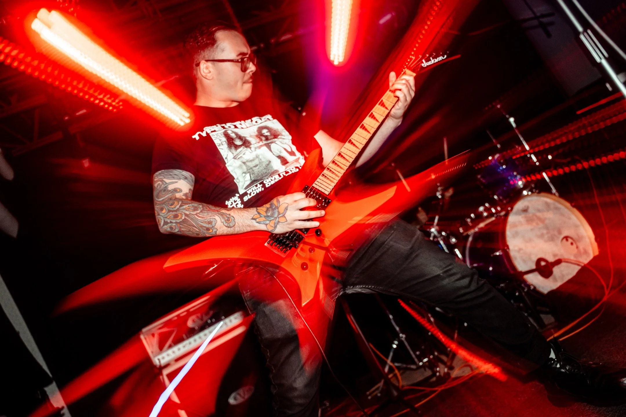 A male guitarist playing an orange electric guitar on stage with red lighting and a drum set in the background.