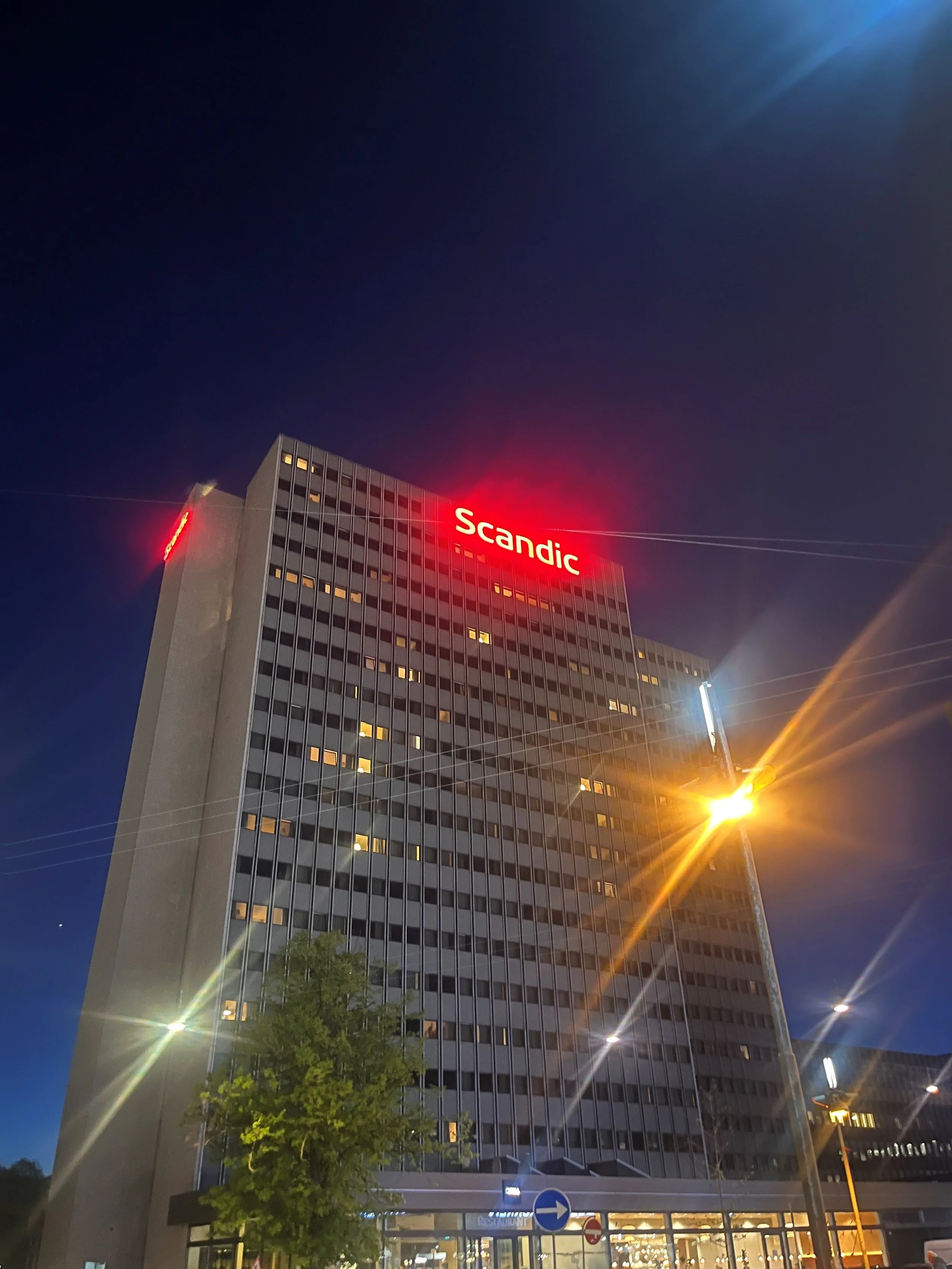 Tall hotel building with "Scandic" in red letters on top, captured at night with illuminated windows and streetlights.