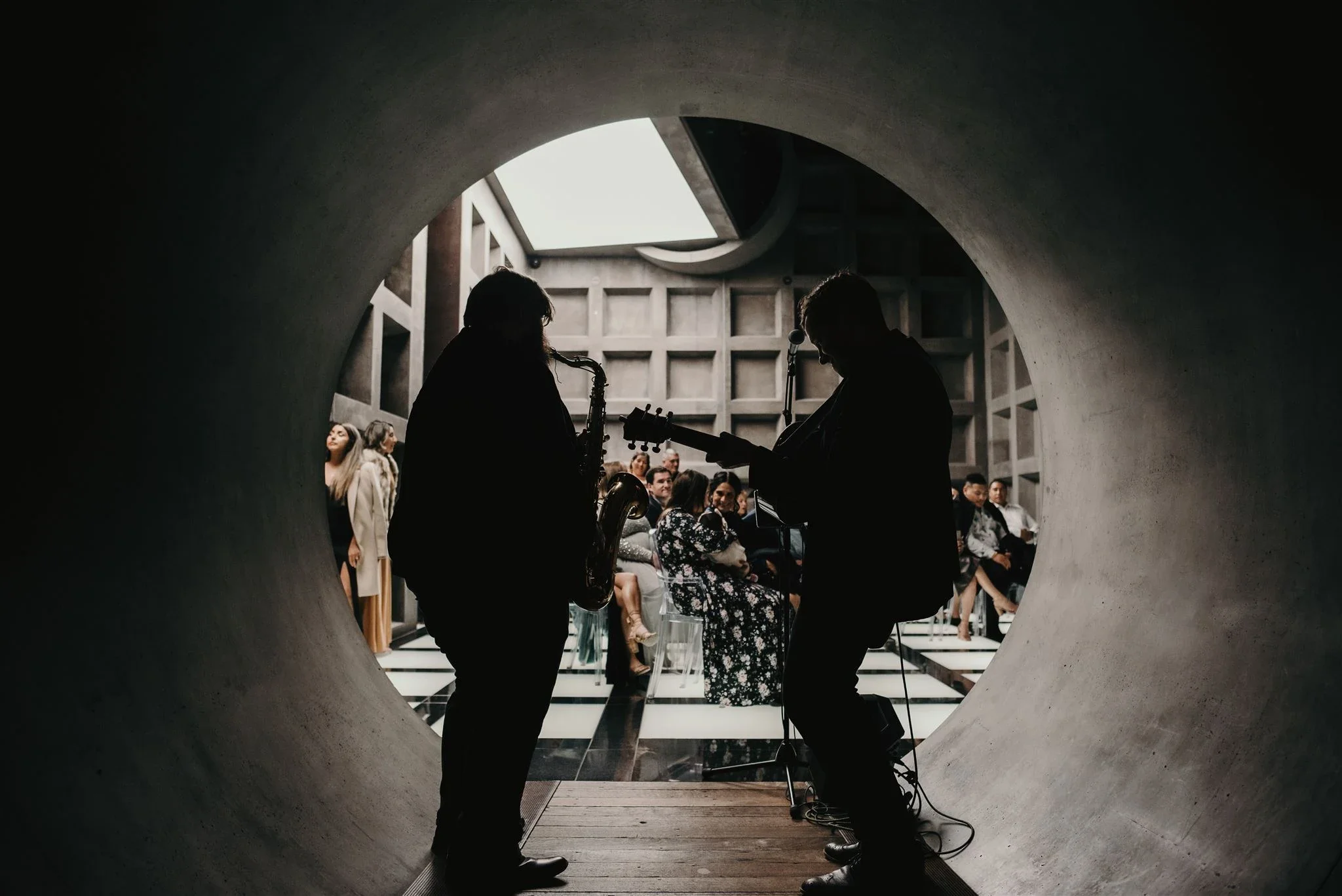 Silhouettes of two musicians, a saxophonist and a guitarist, performing inside a circular tunnel with an audience in the background.