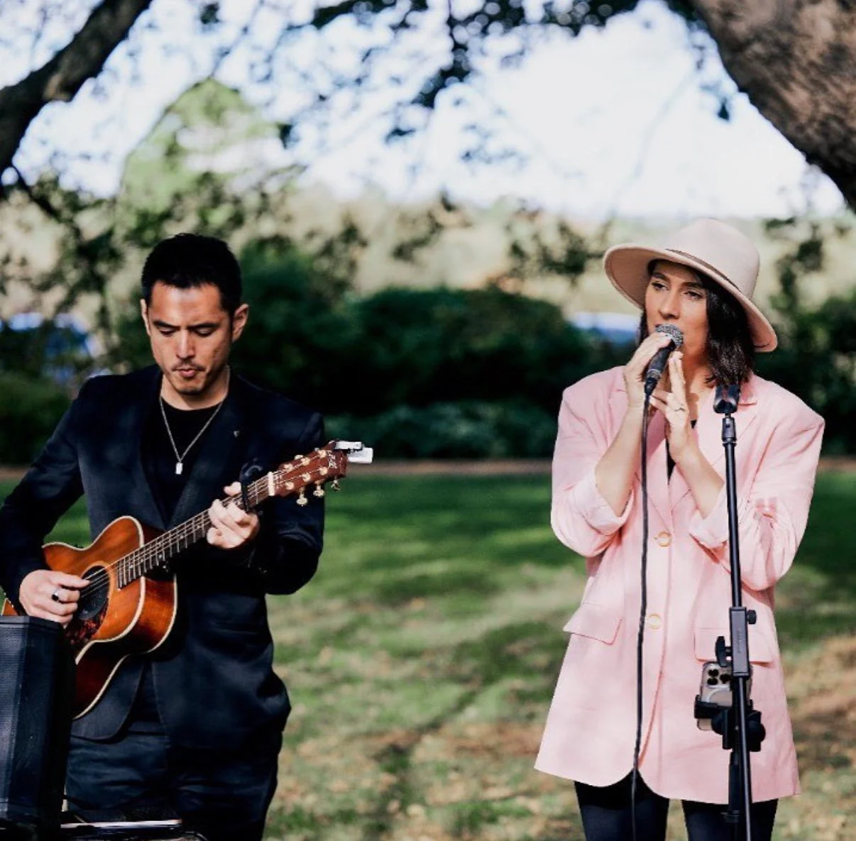 A man playing an acoustic guitar and a woman singing into a microphone outdoors under a tree with greenery in the background.