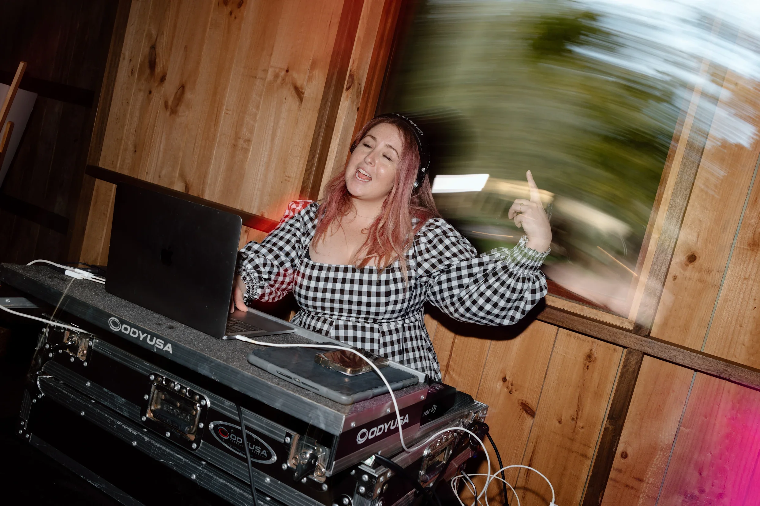 Female DJ with headphones DJing in a wood-paneled room, wearing a black and white checkered dress, with her eyes closed and her mouth open, dancing or singing, with DJ equipment and a laptop in front of her.