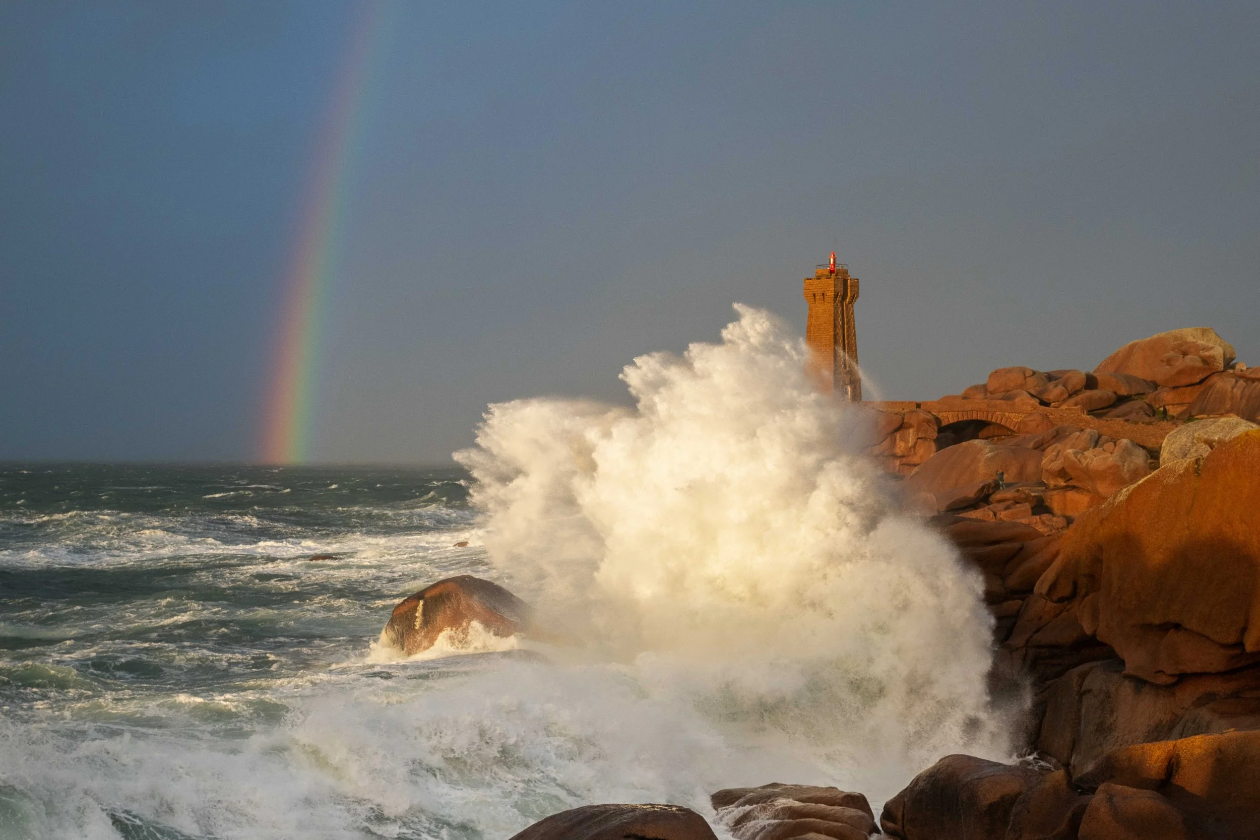 Vagues s'écrasant contre des rochers avec le phare Mean Ruz en arrière-plan et un arc-en-ciel dans le ciel nuageux au coucher du soleil.