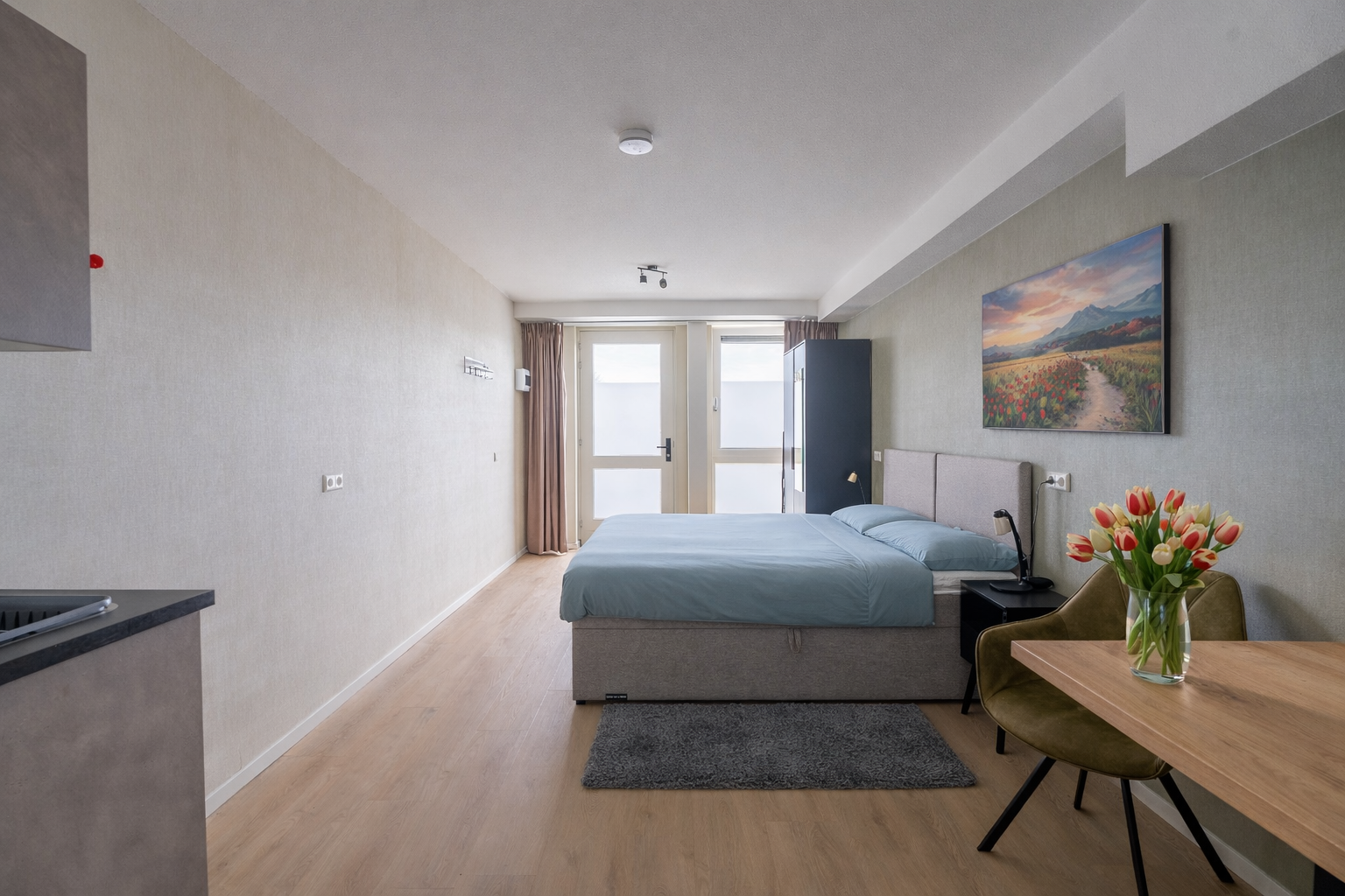 A bedroom with a gray bed, a black side table, a vase of tulips on a wooden table, a painting of a landscape, and a sliding glass door with beige curtains.