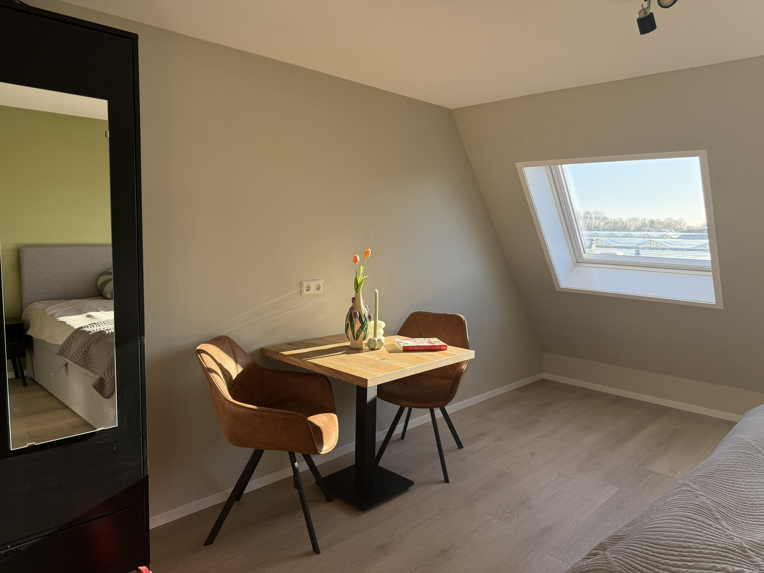 A small room with a slanted ceiling, a rectangular window, a wooden table with two brown chairs, a vase with tulips, a book, and a bed visible through an open door.