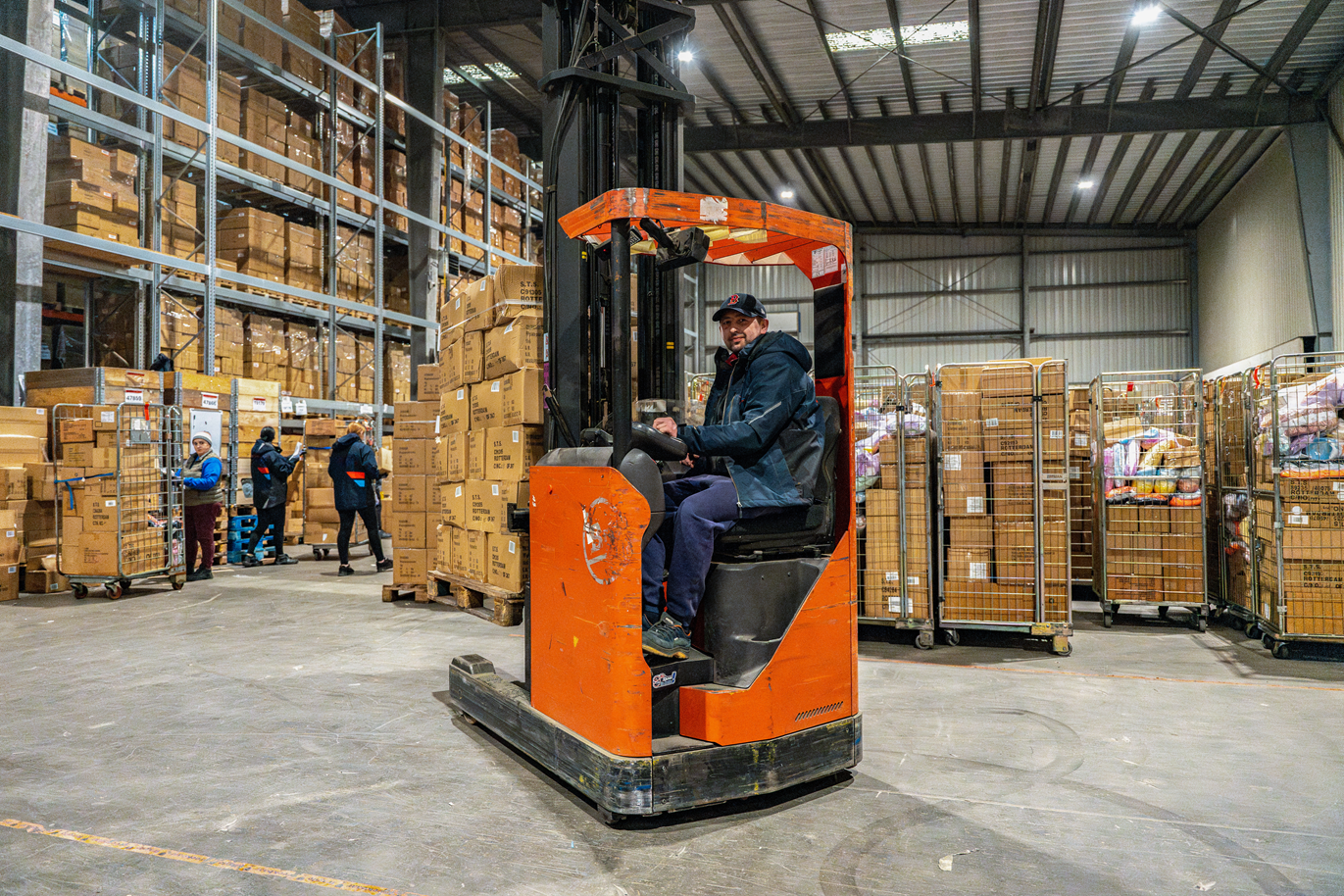 A warehouse worker operating an orange forklift among tall shelves stacked with cardboard boxes and containers, with other workers in the background handling boxes.