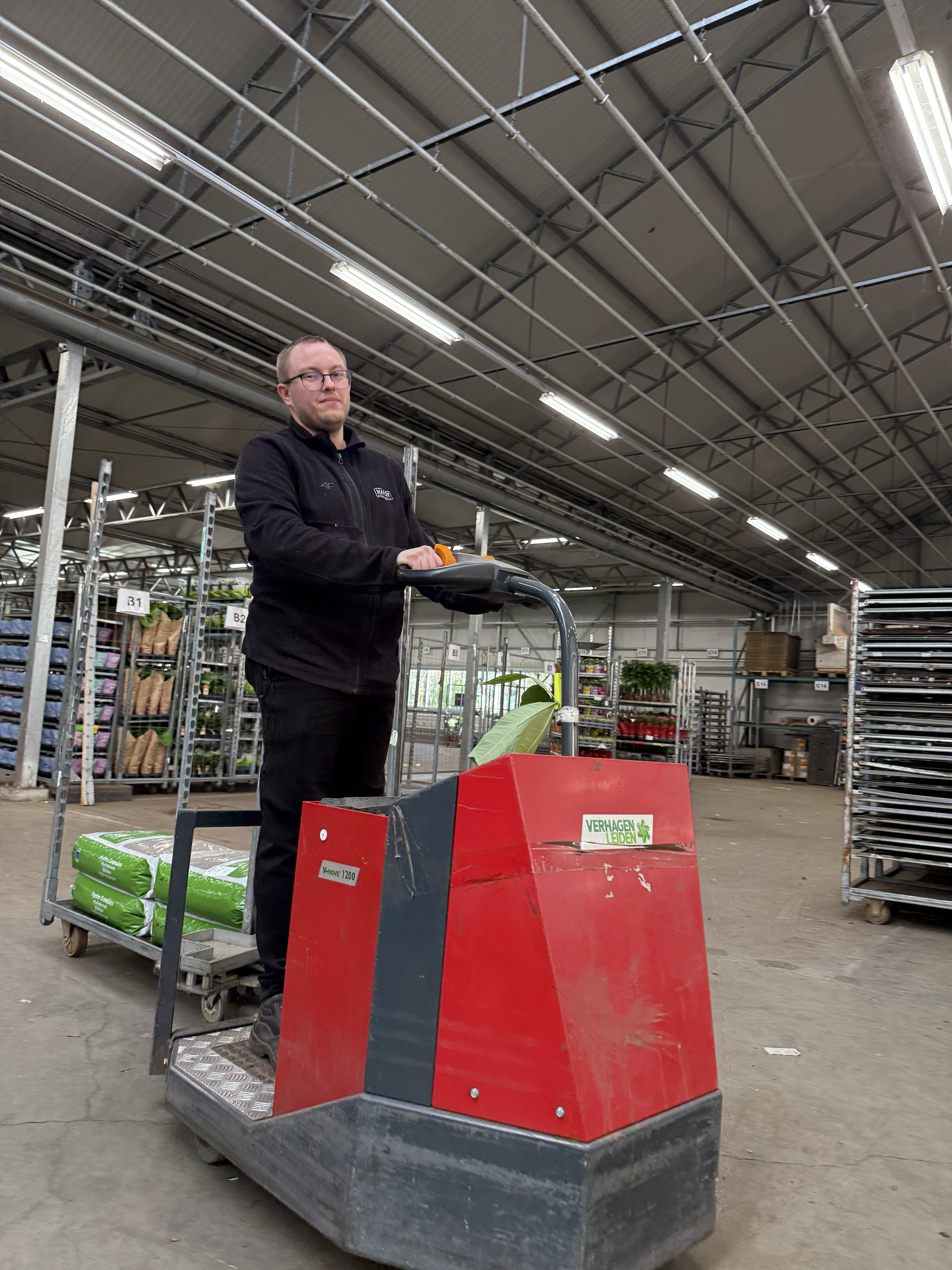 A man operating a red walk-behind trowel in a warehouse or garden center with shelves of plants and supplies.
